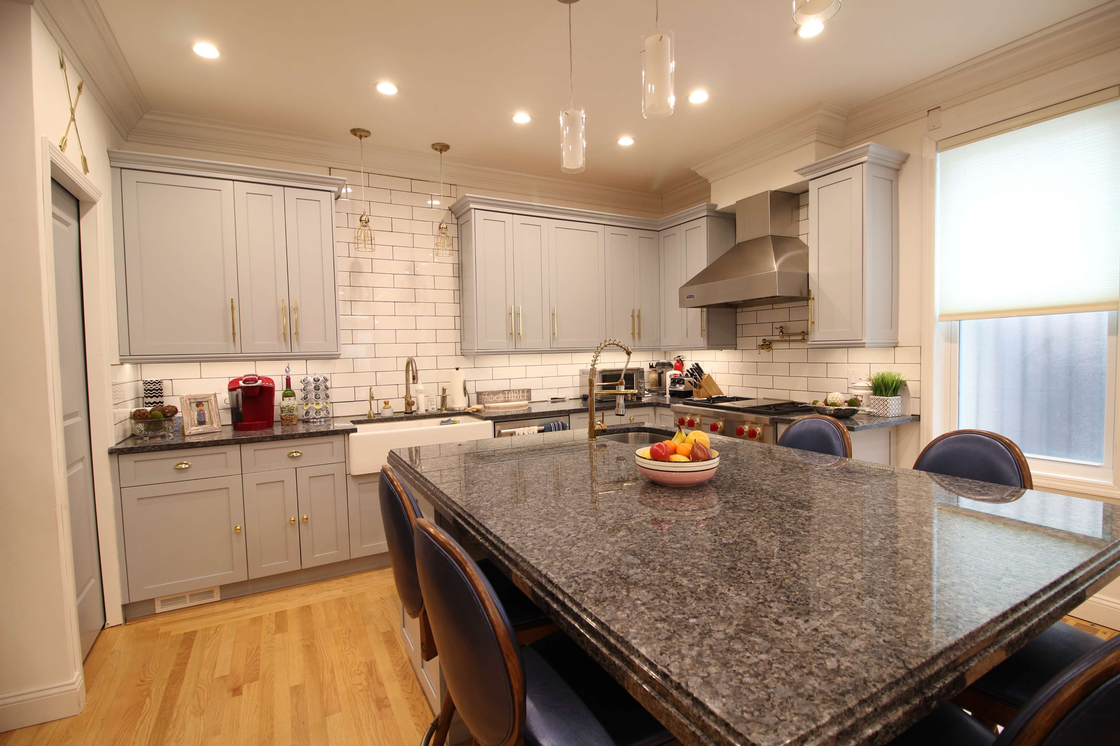 A modern kitchen featuring gray cabinets, a large granite island, stainless steel appliances, and a white tile backsplash.