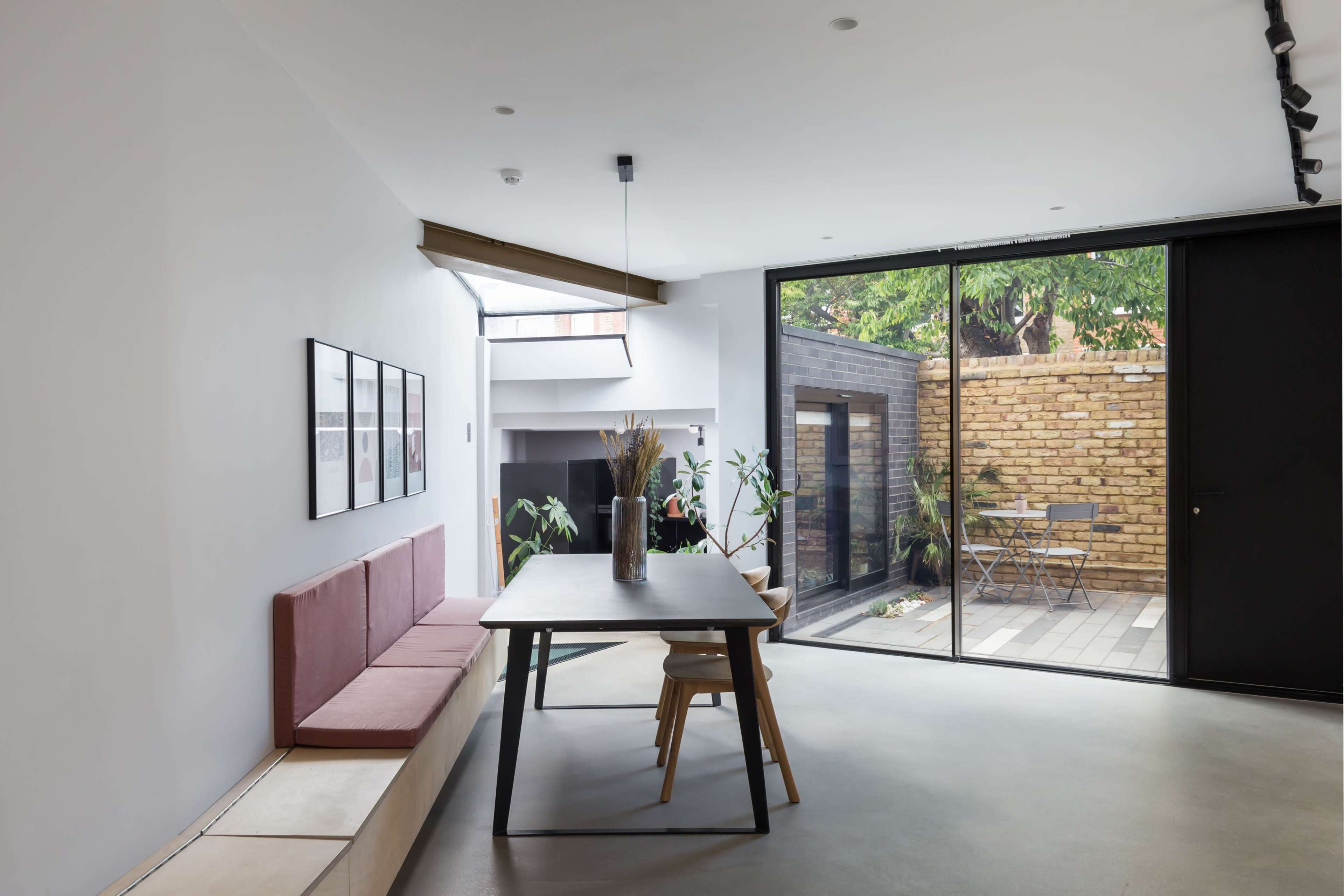 A modern dining area with a large table, minimalist decor, and large sliding glass doors opening to a small outdoor patio surrounded by greenery.