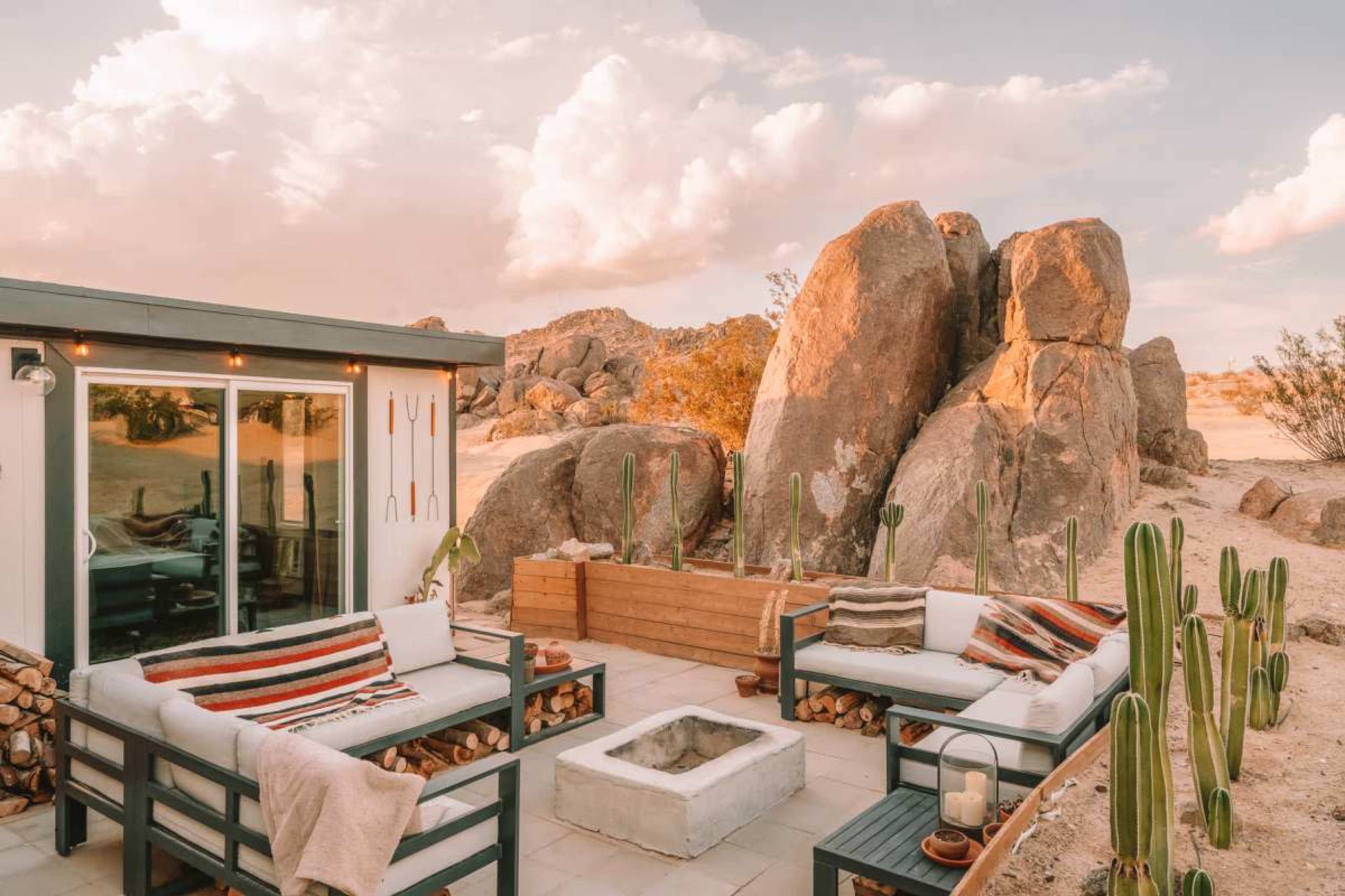 The image shows a modern outdoor living space with couches, a fire pit, and large boulders in the background.