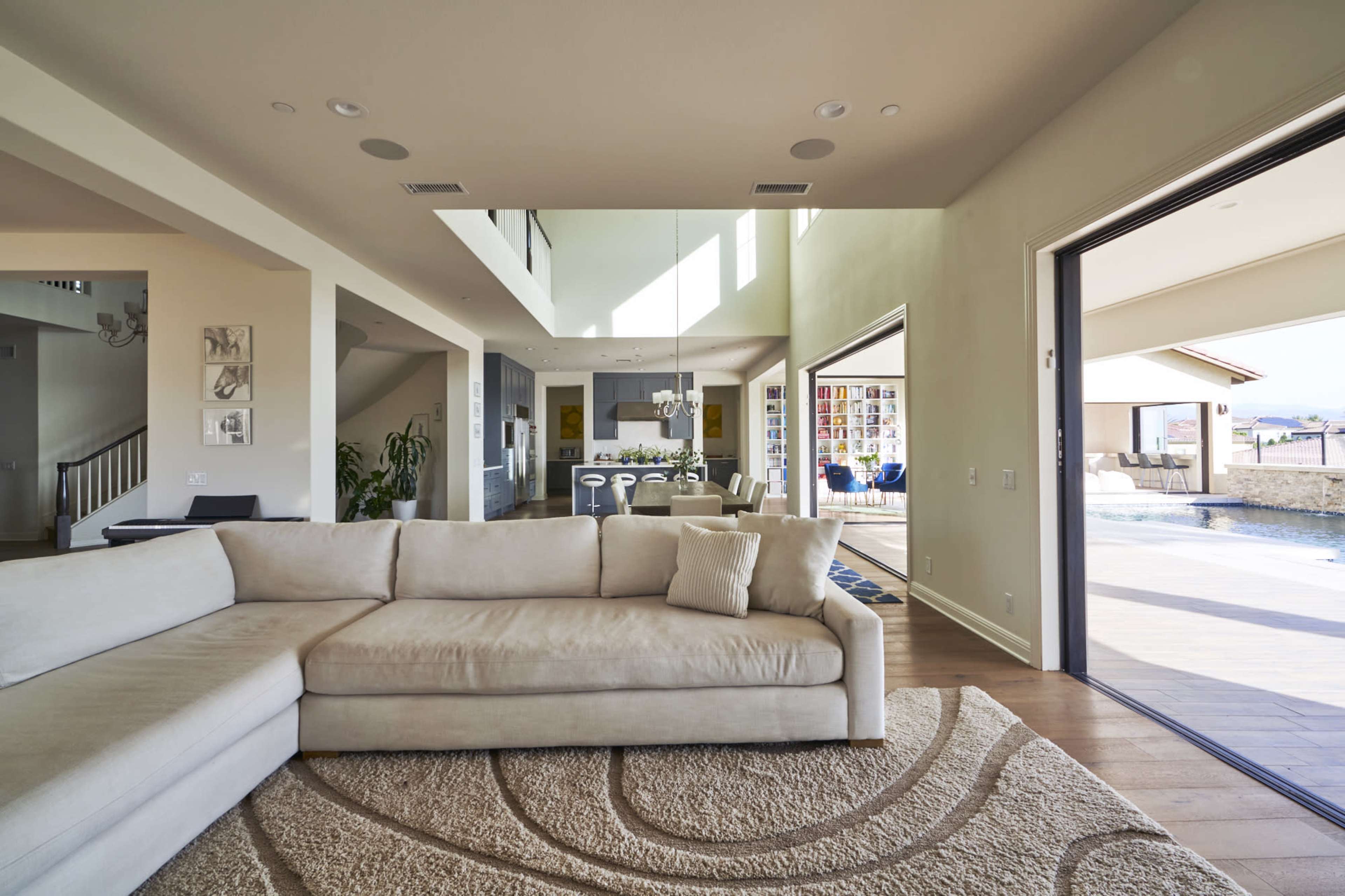 A spacious living room with a light-colored sectional sofa, leading to an open dining area and a view of an outdoor pool.