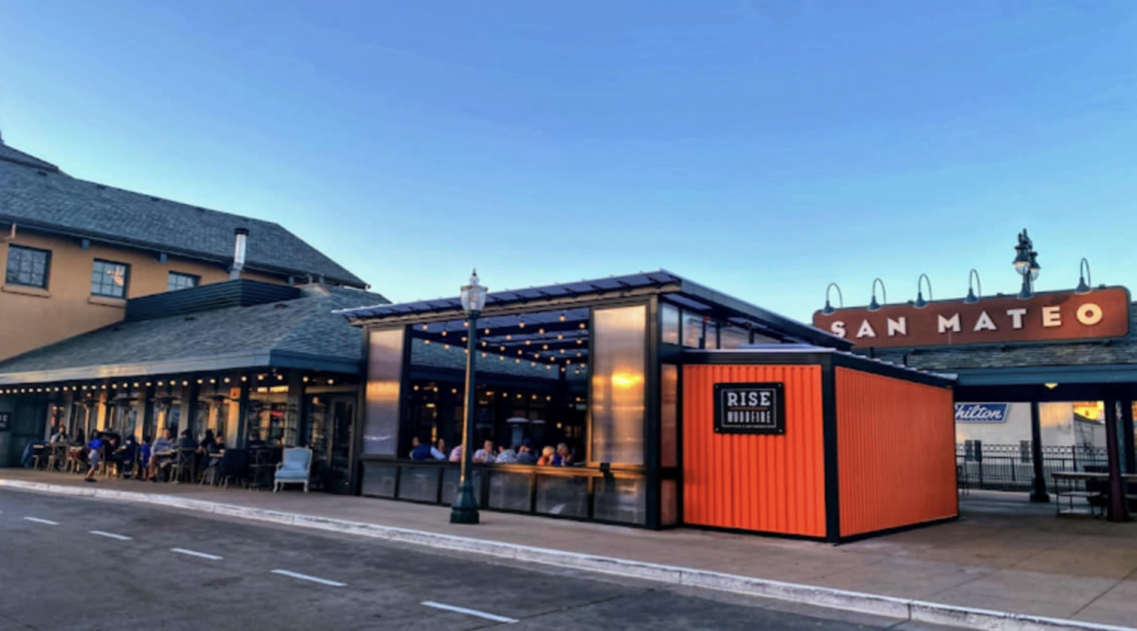 A modern restaurant with an outdoor seating area, situated next to a historic building and a sign that reads "SAN MATEO."
