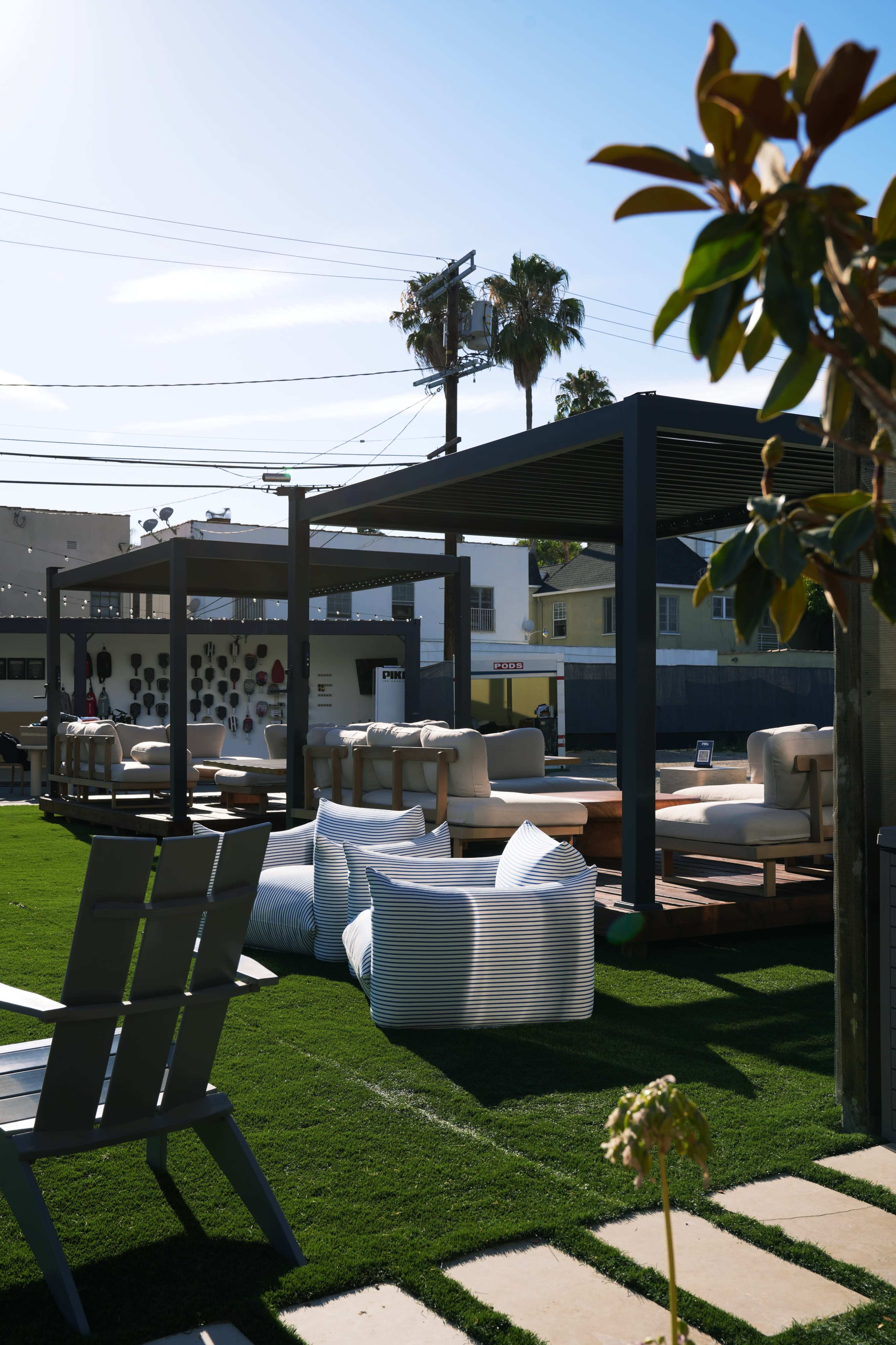 The image shows a seating area with wooden platforms and white cushions on a grassy lawn, surrounded by palm trees and buildings in the background.