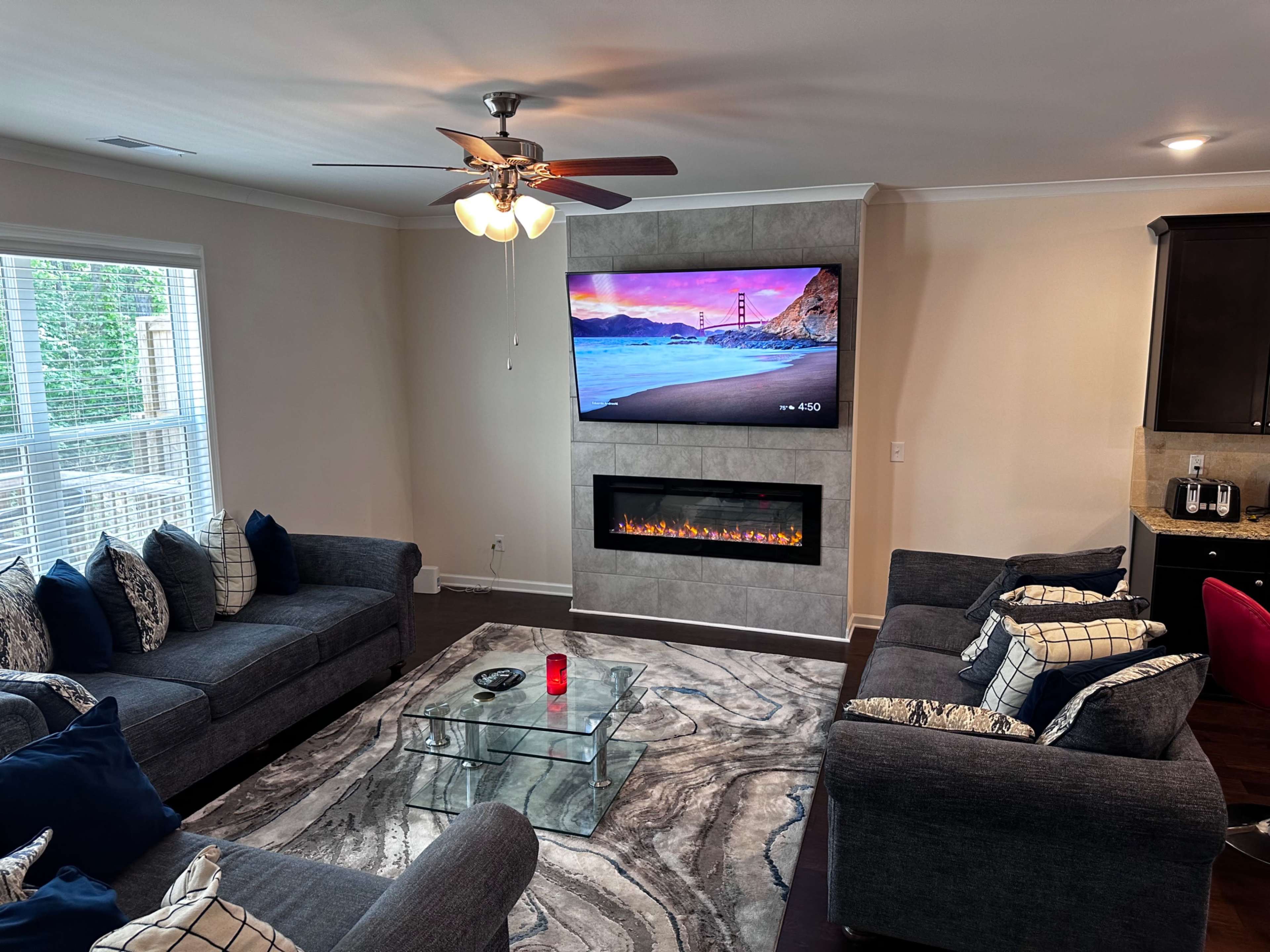 A modern living room featuring two gray sofas, a glass coffee table, a mounted television, and a fireplace beneath the TV, all set against light-colored walls and hardwood flooring.