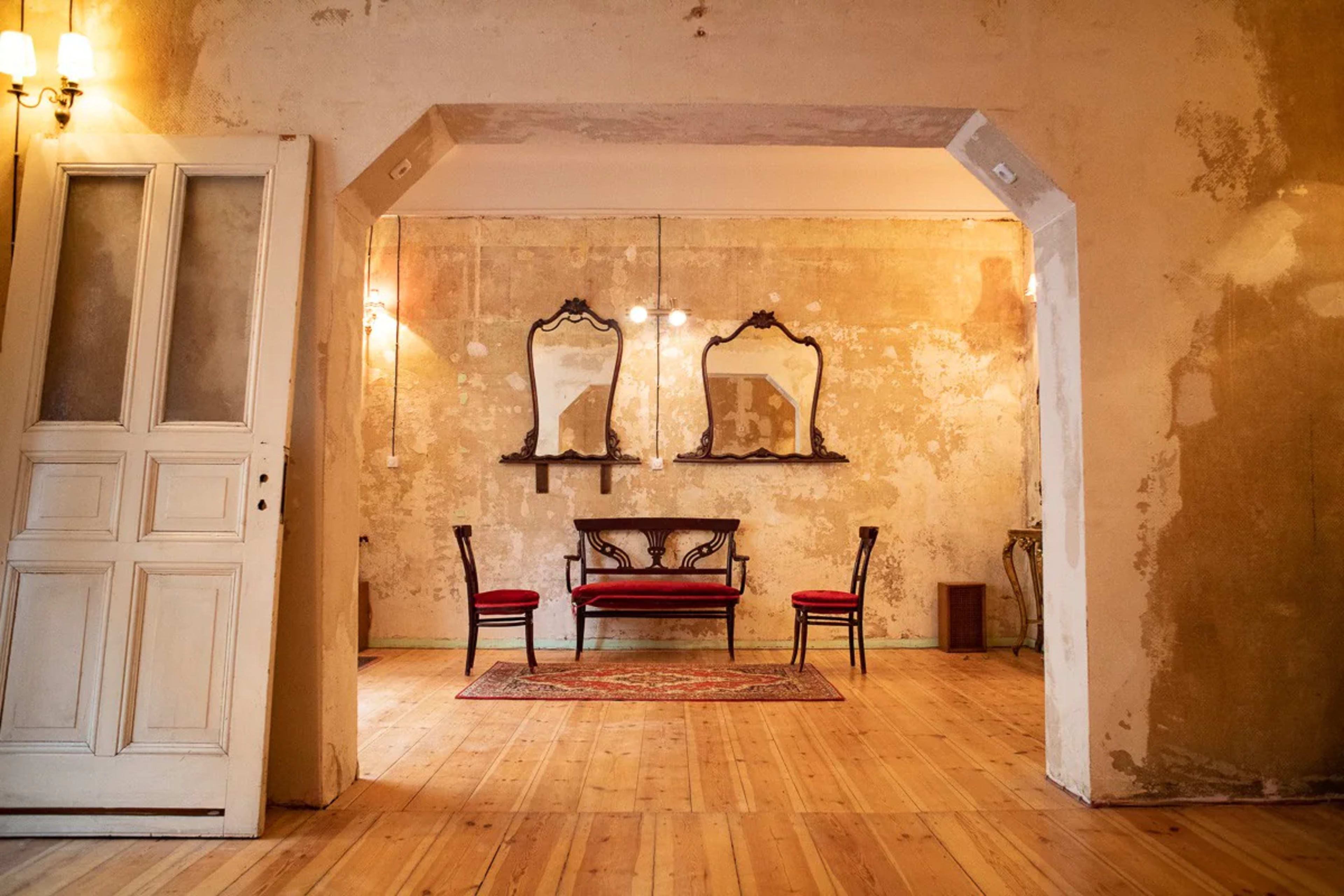 A room with a wooden floor, two red chairs, a central ornate bench, and three framed mirrors on the wall, all set against a peeling plaster backdrop.
