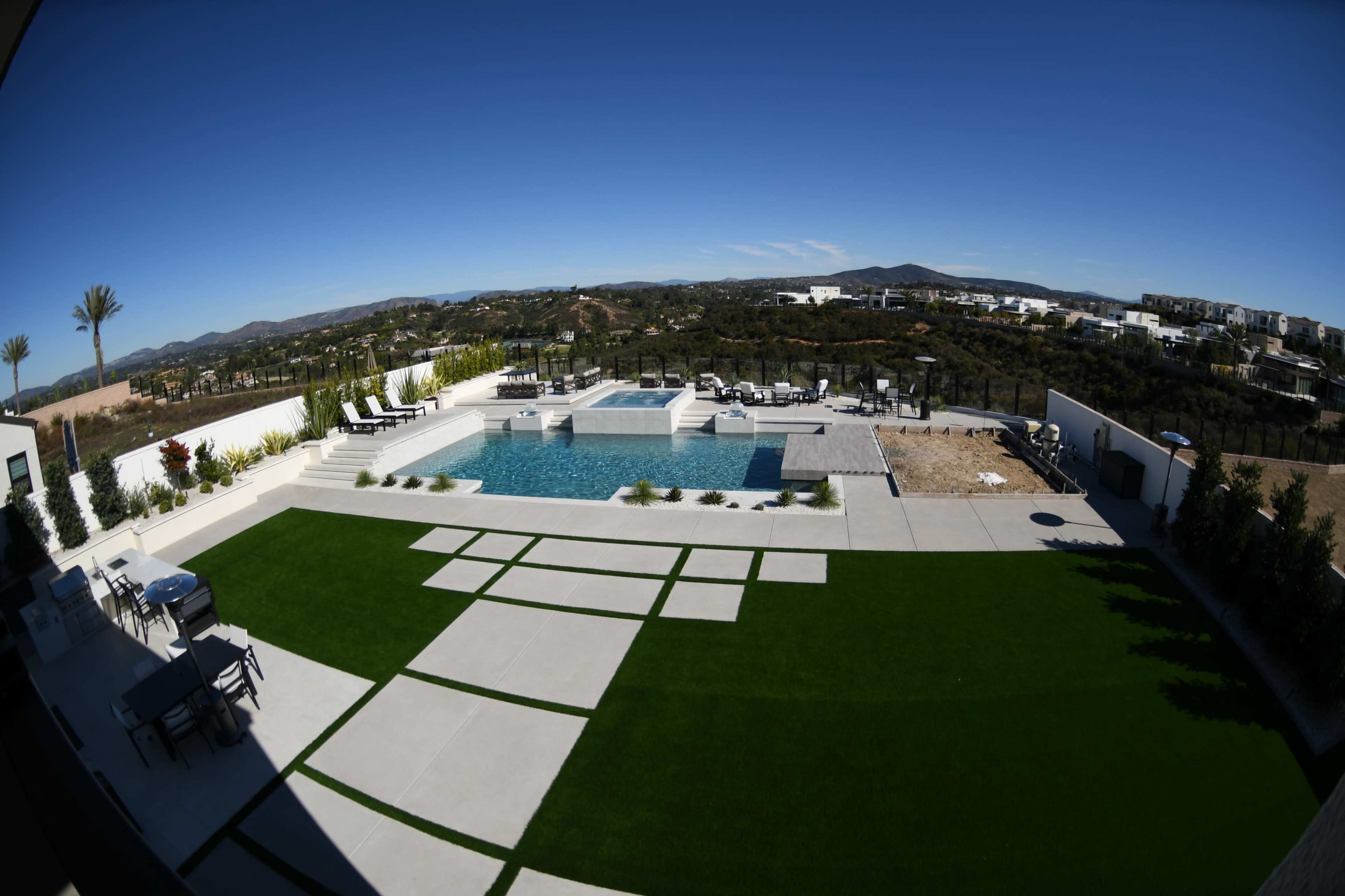 A large outdoor area featuring a swimming pool, lounge chairs, and a manicured lawn, set against a backdrop of rolling hills and clear blue skies.