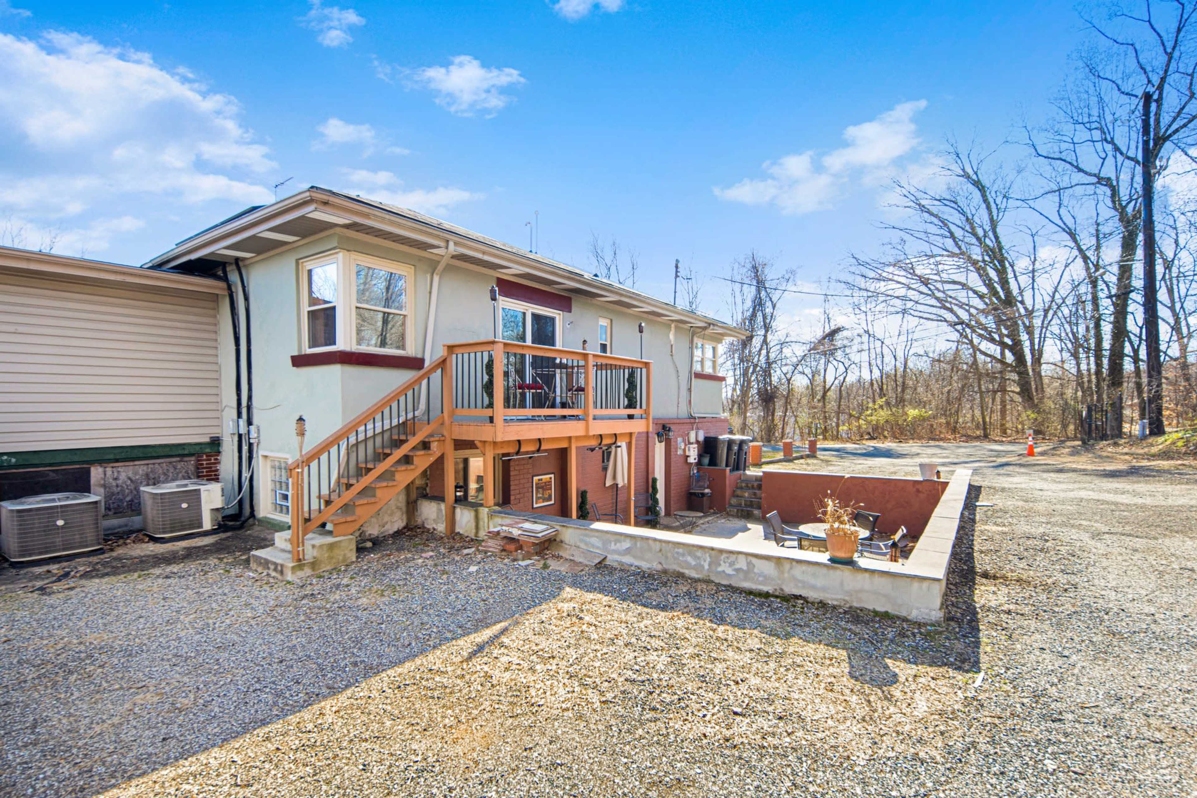 A two-story house with a wooden stairway leading to a deck on the upper level, surrounded by a gravel area and bare trees in the background.