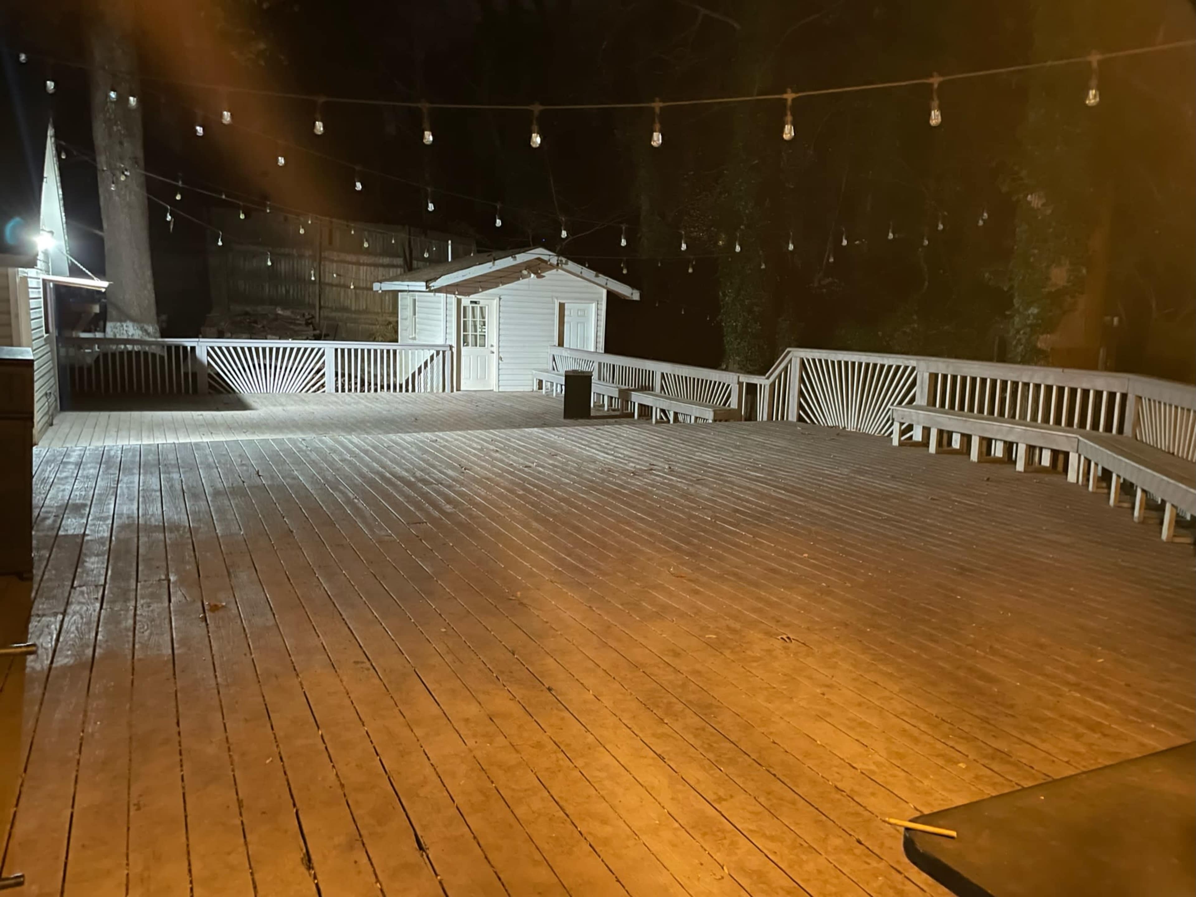 A large wooden deck is illuminated by string lights and features a small building and benches along the perimeter.