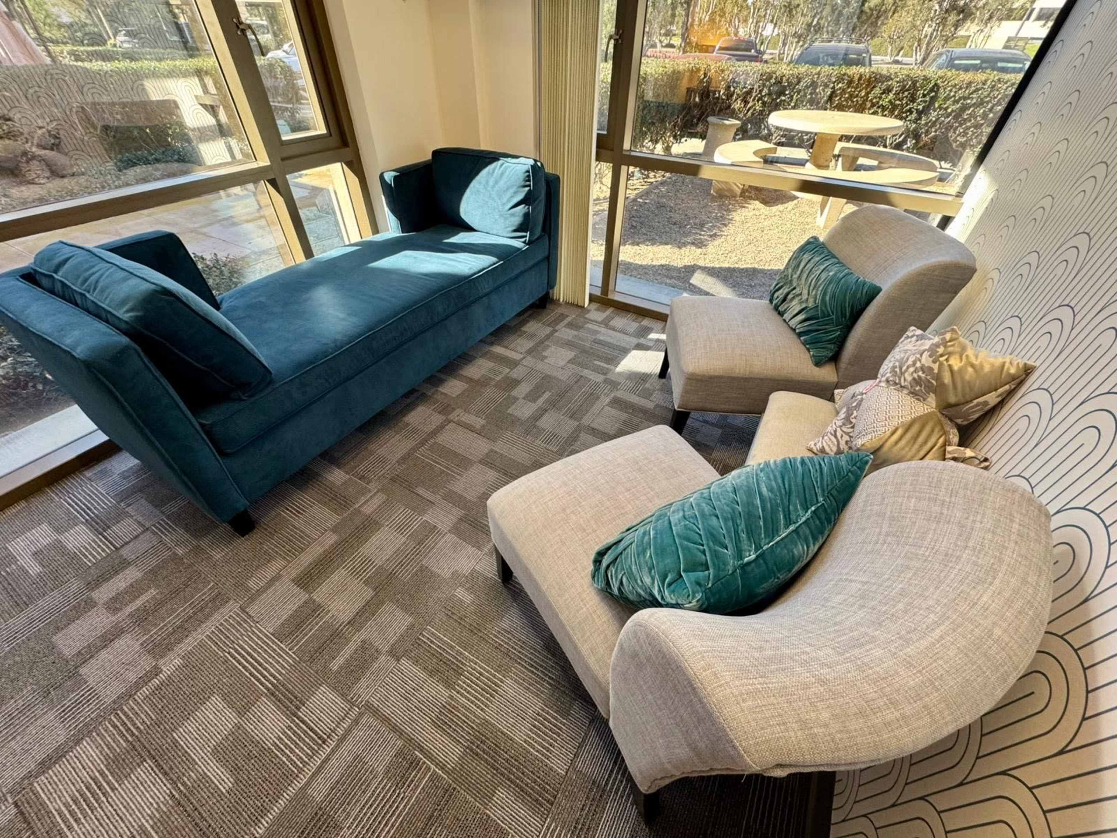 The image shows a cozy seating area with a teal couch and two light gray armchairs, positioned near large windows that let in natural light.