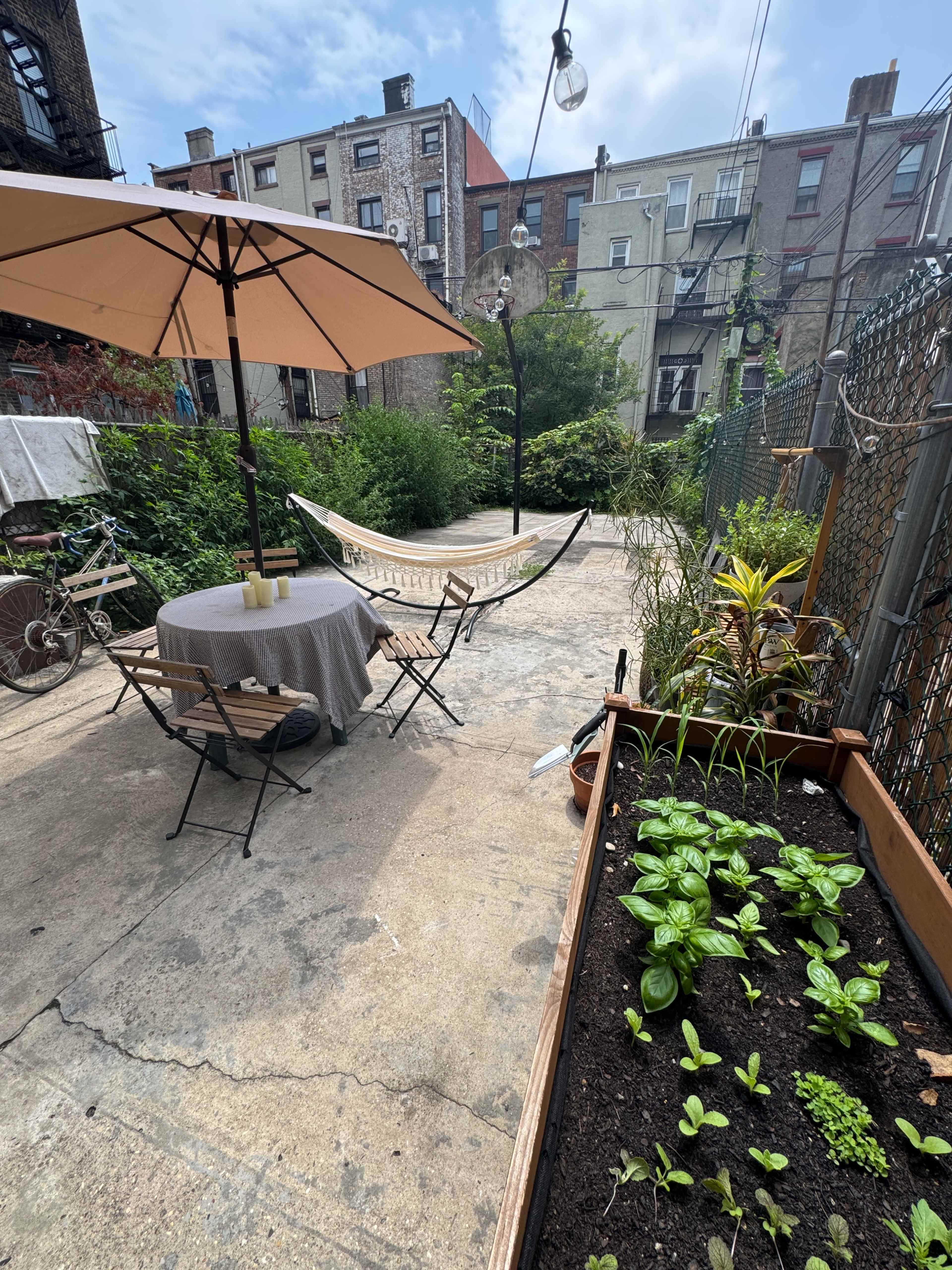 The image shows a backyard garden with a table and chairs under an umbrella, a hammock, and a planter containing young vegetables, surrounded by tall greenery and urban buildings.