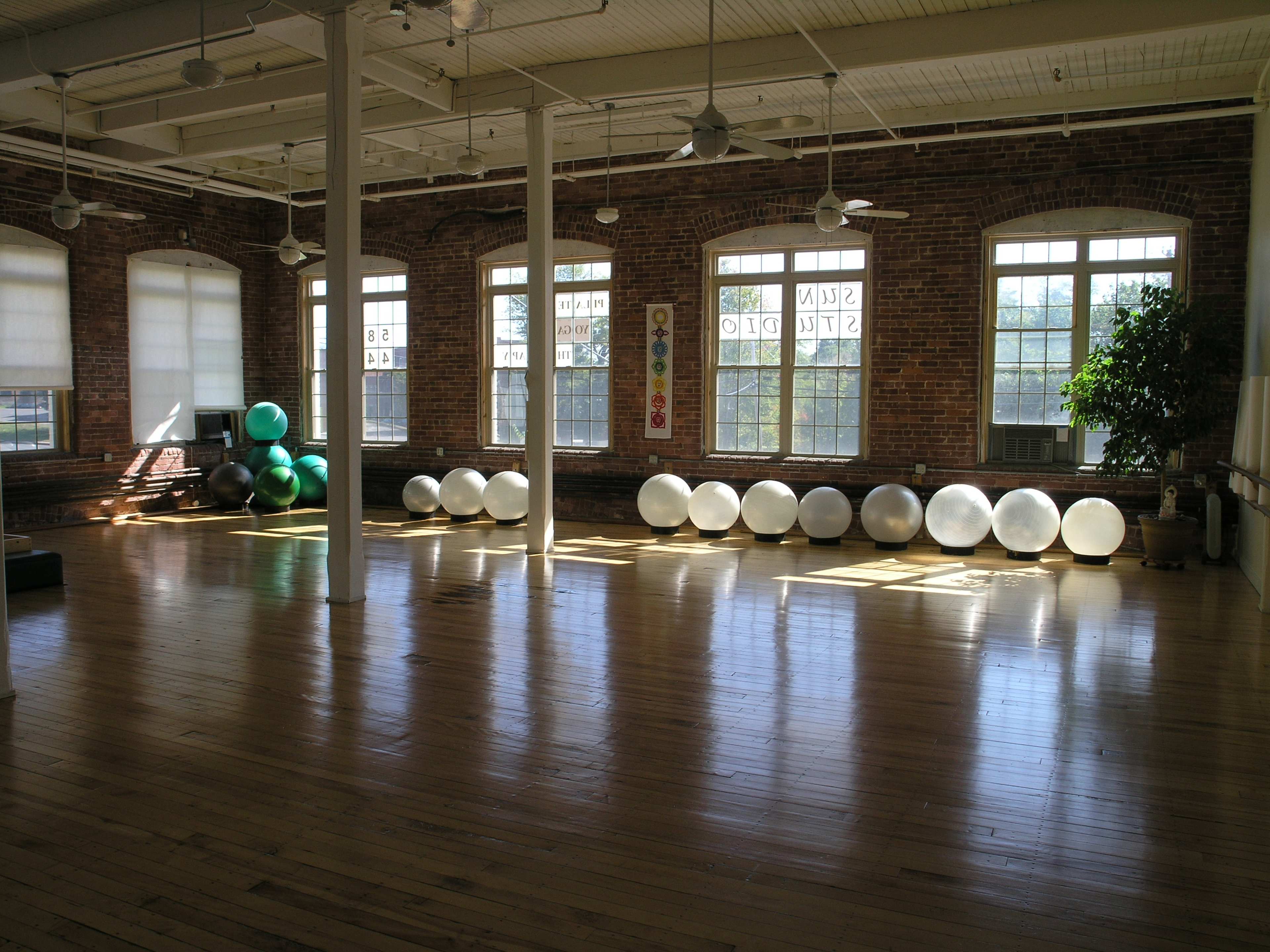 An open room with wooden floors features large exercise balls arranged along a wall, illuminated by sunlight streaming through multiple windows.