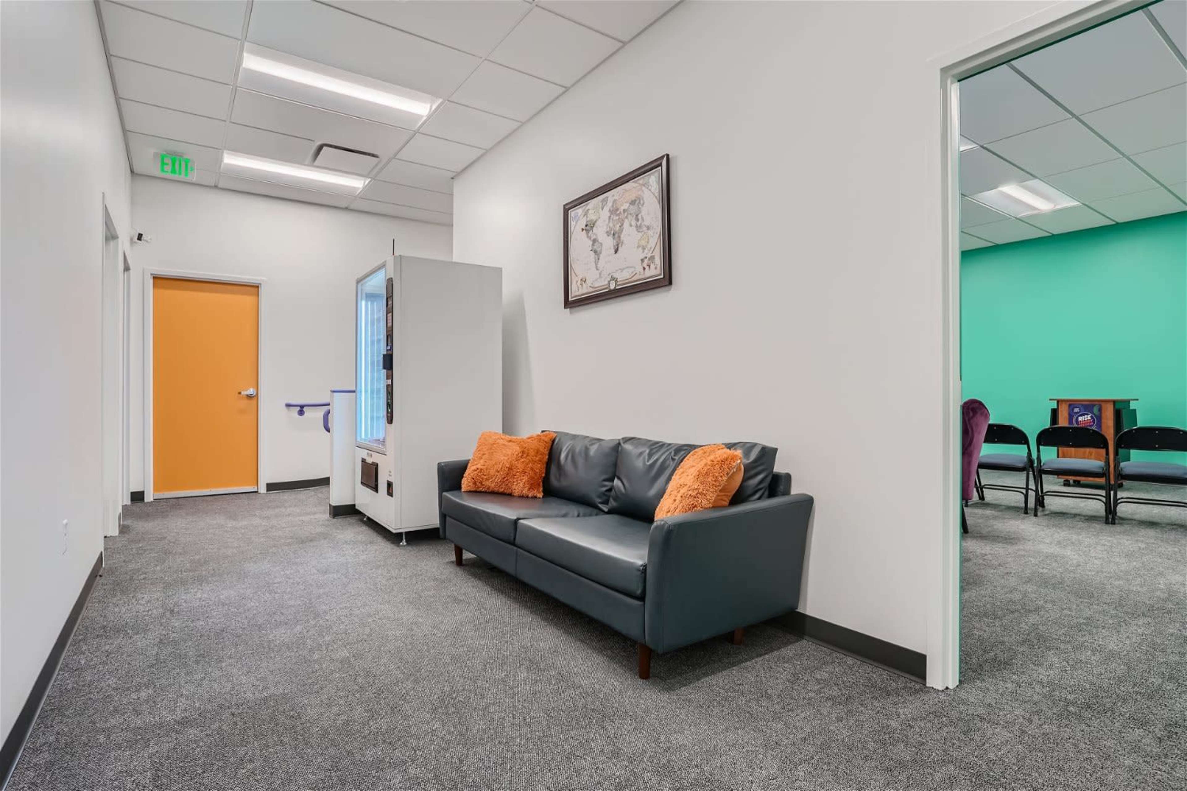 A hallway with a black couch, a vending machine, an orange door, and a green room visible in the background.