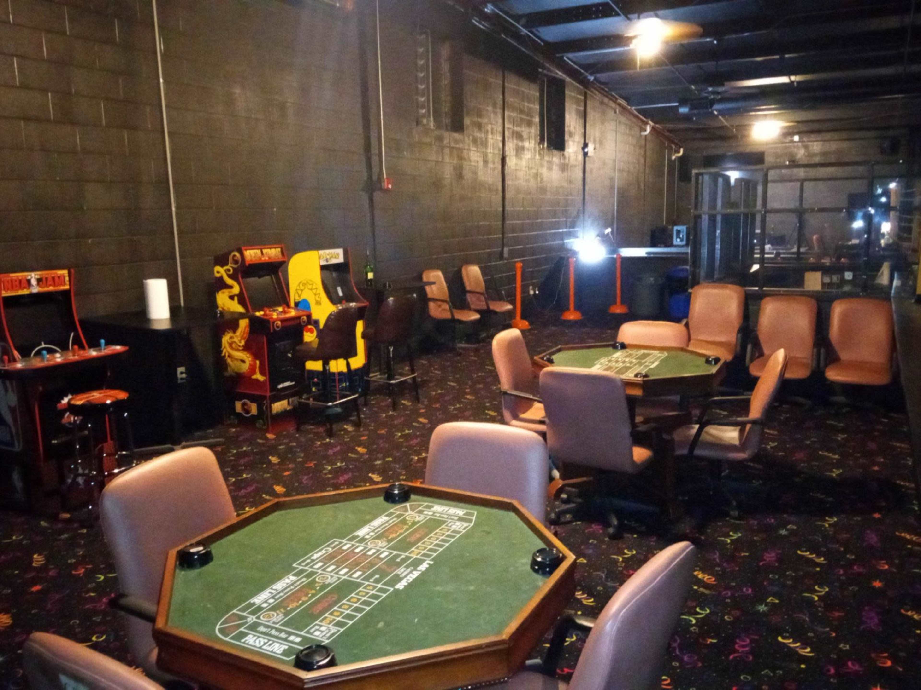 The image shows a dimly lit gaming area with several green felt gaming tables, a few arcade machines, and empty brown chairs arranged around the tables.