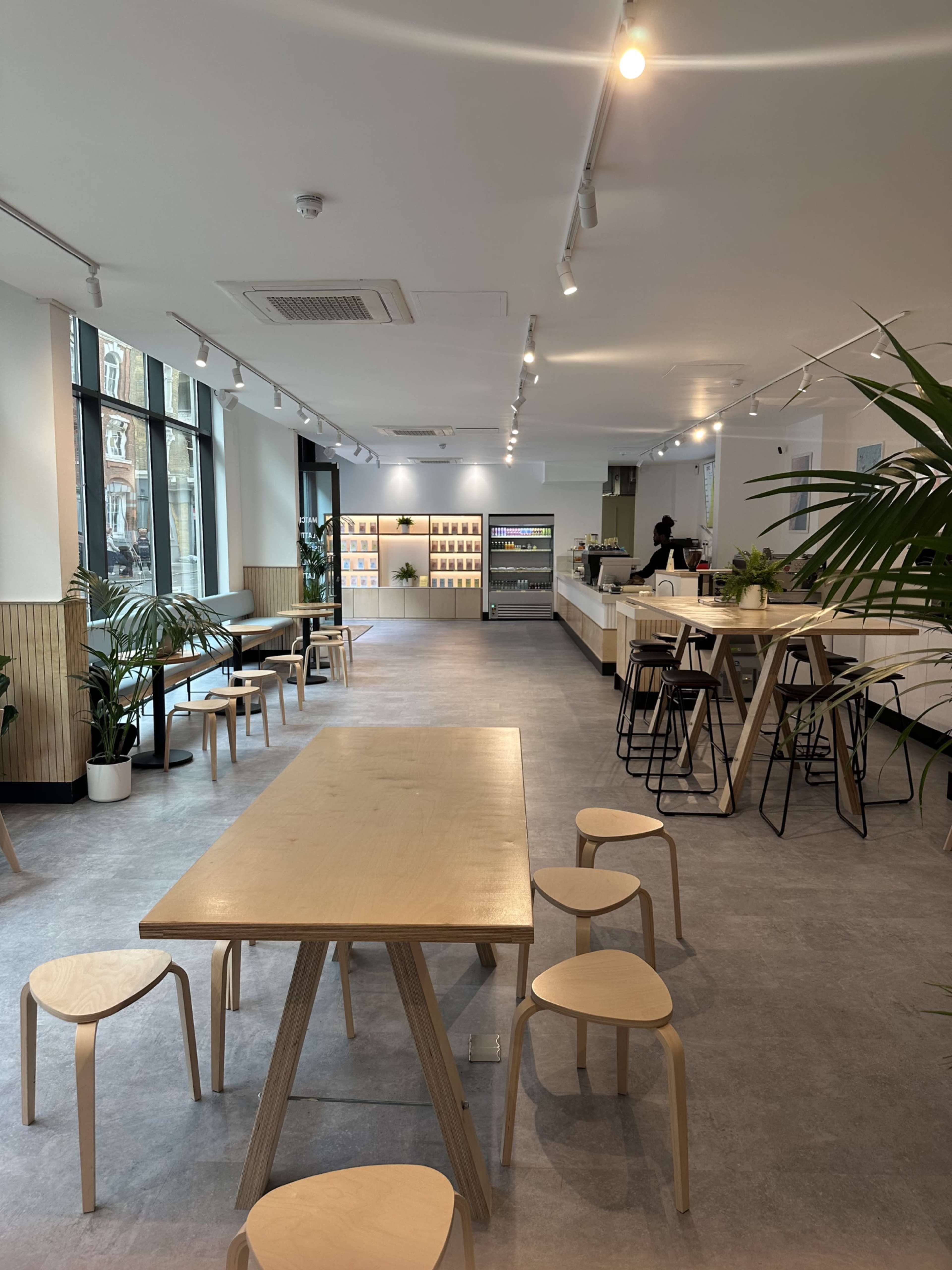 The image shows a modern café interior with a light color scheme, featuring wooden tables, minimalist seating, large windows, and a counter area.
