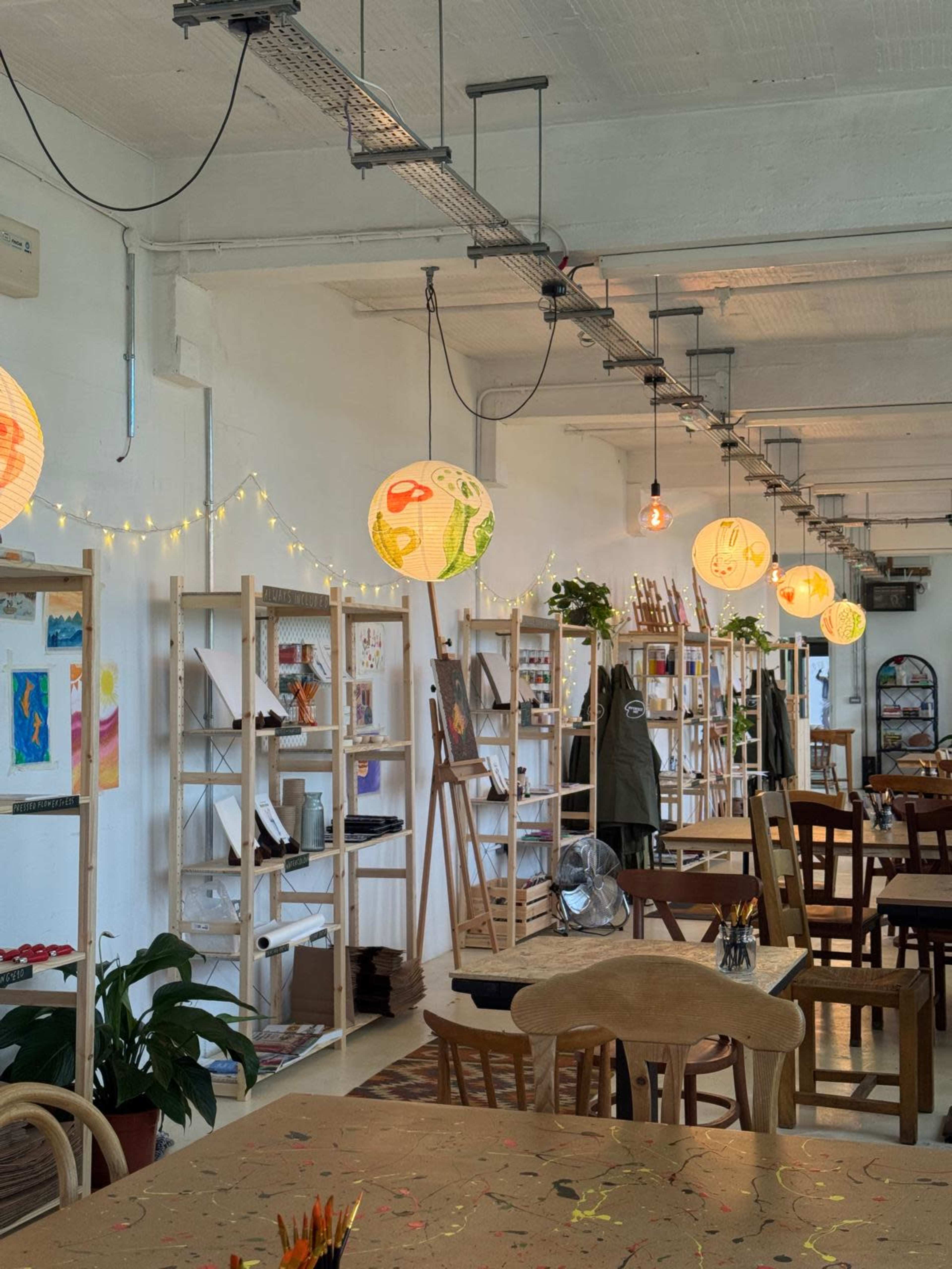 The image shows an art studio with shelves displaying artwork, tables for creative work, and hanging decorative lanterns.