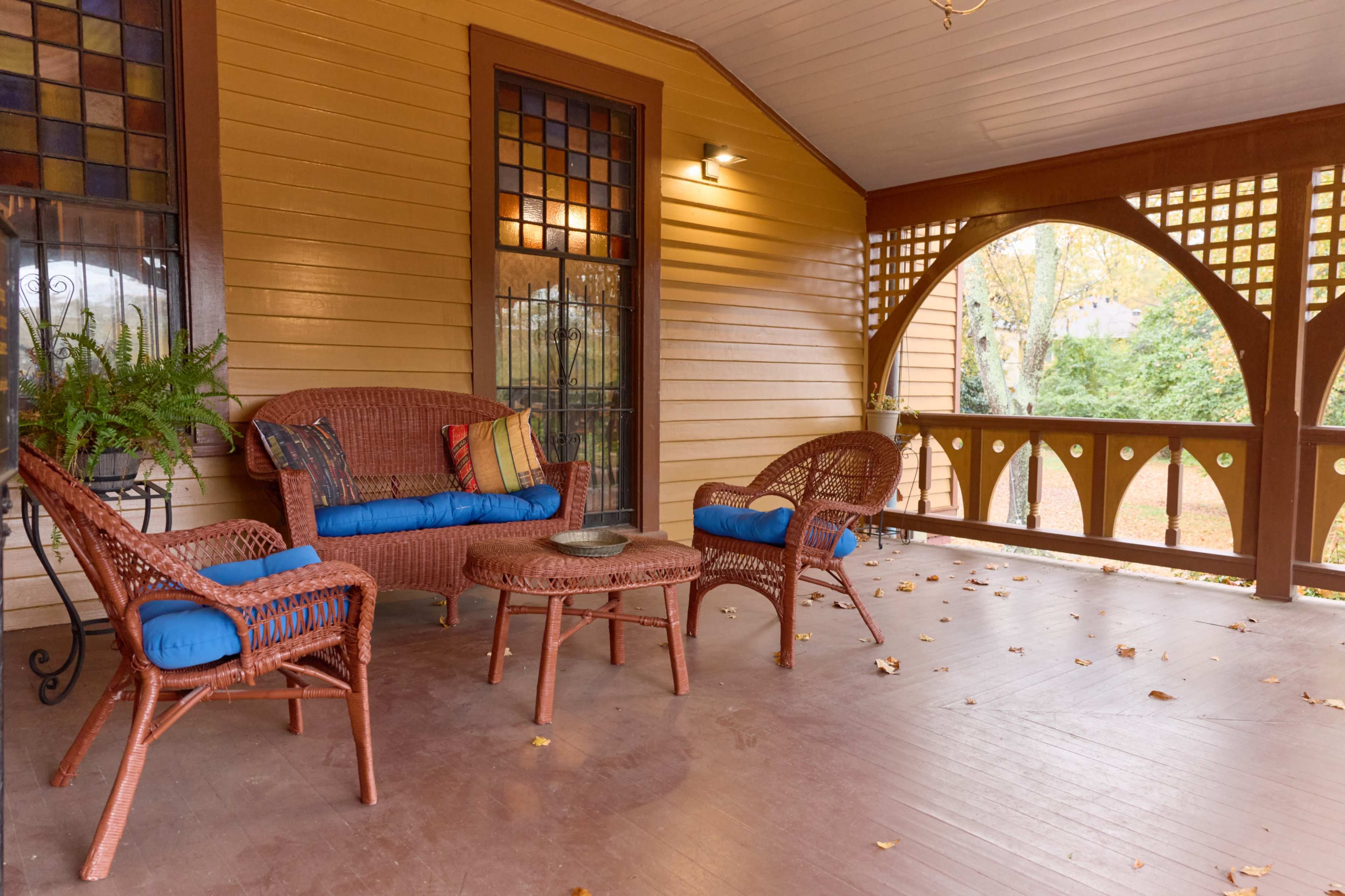 The image shows a porch with wicker furniture, featuring a sofa and chairs with blue cushions, and a circular table, surrounded by wooden paneling and stained glass windows.