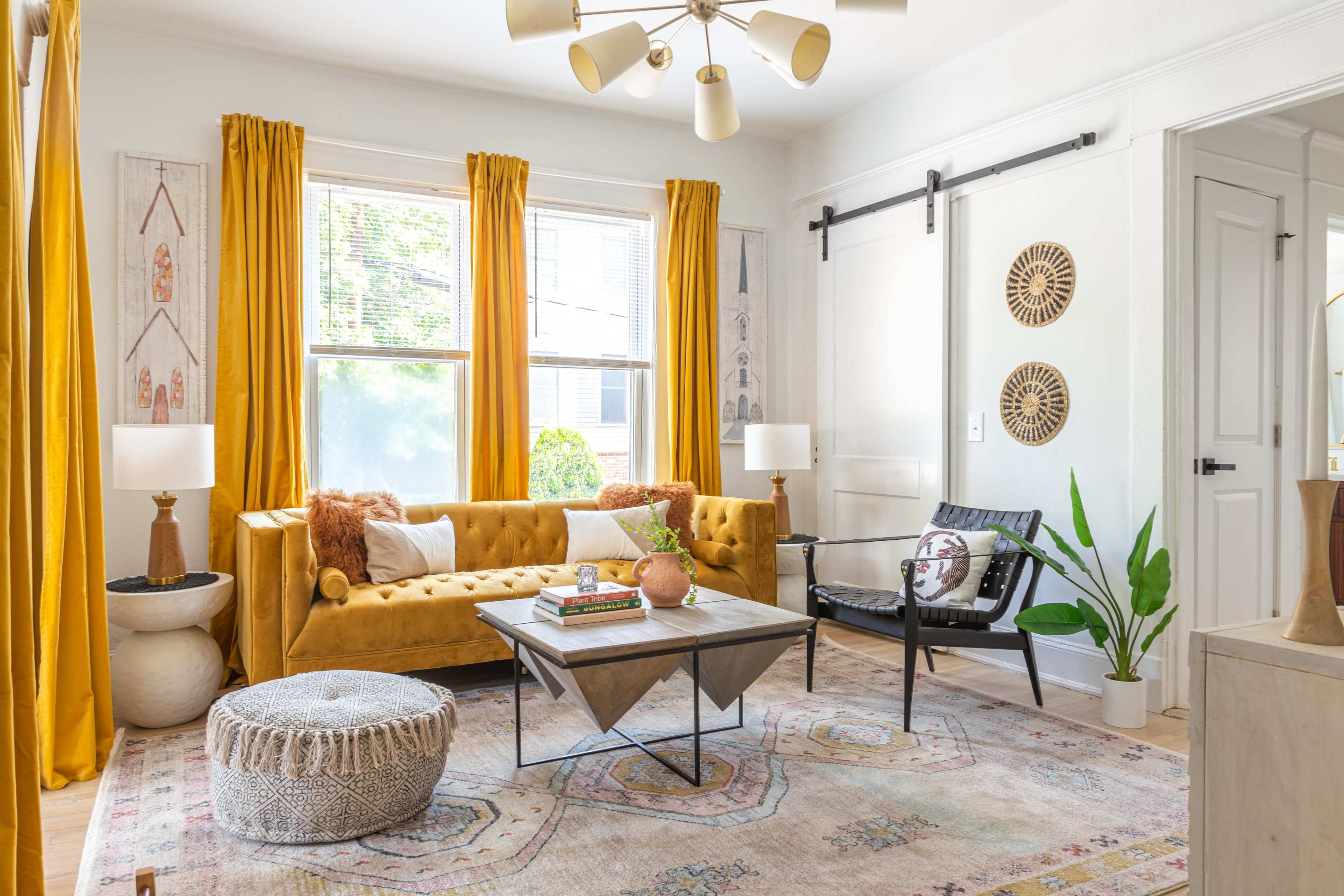 The living room features a mustard yellow sofa, a black chair, and a coffee table on a patterned rug, surrounded by large windows with yellow curtains.