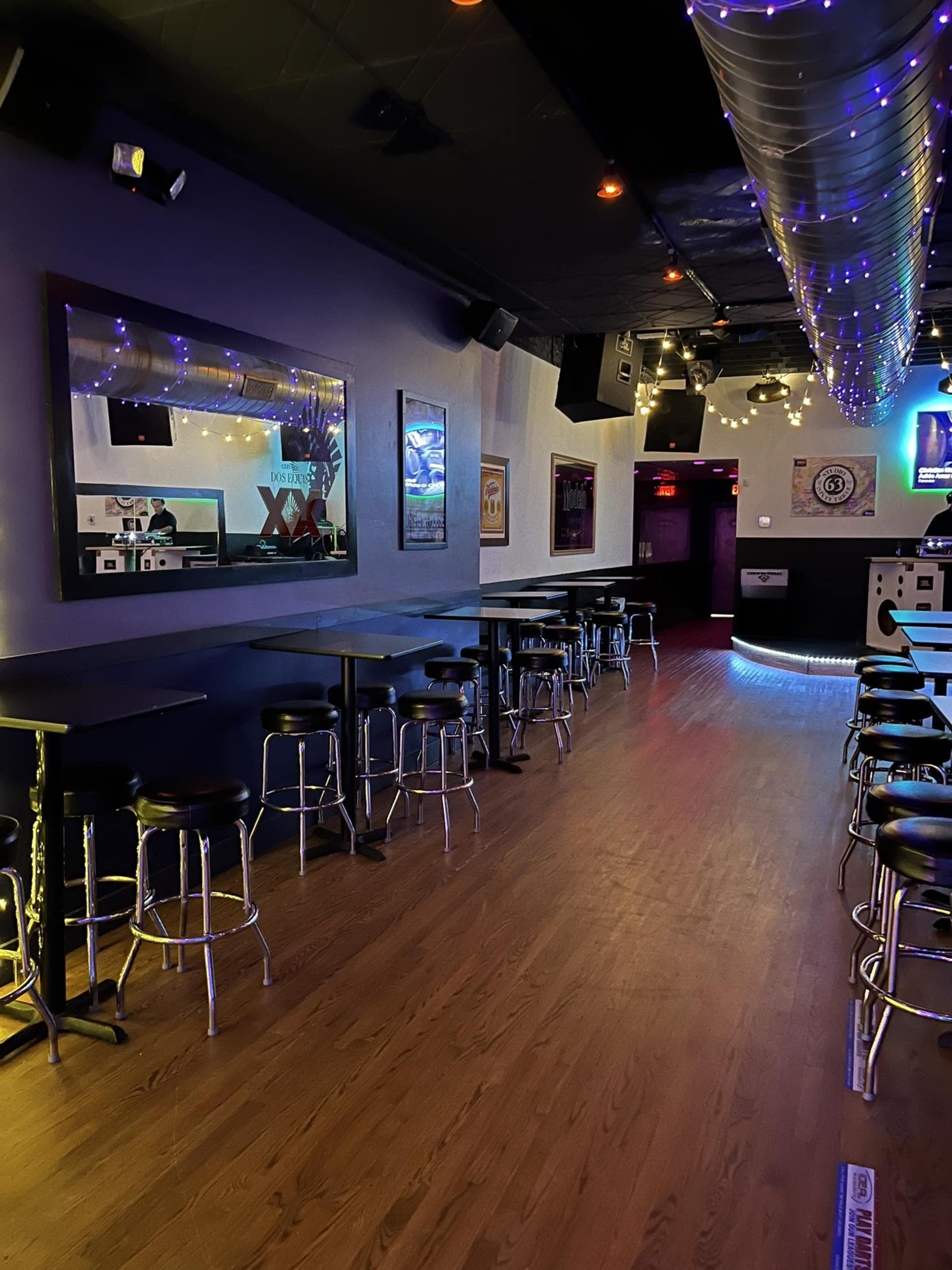 An empty bar interior with high-top tables and chairs, illuminated by soft lighting and decorated with mirrors and wall art.