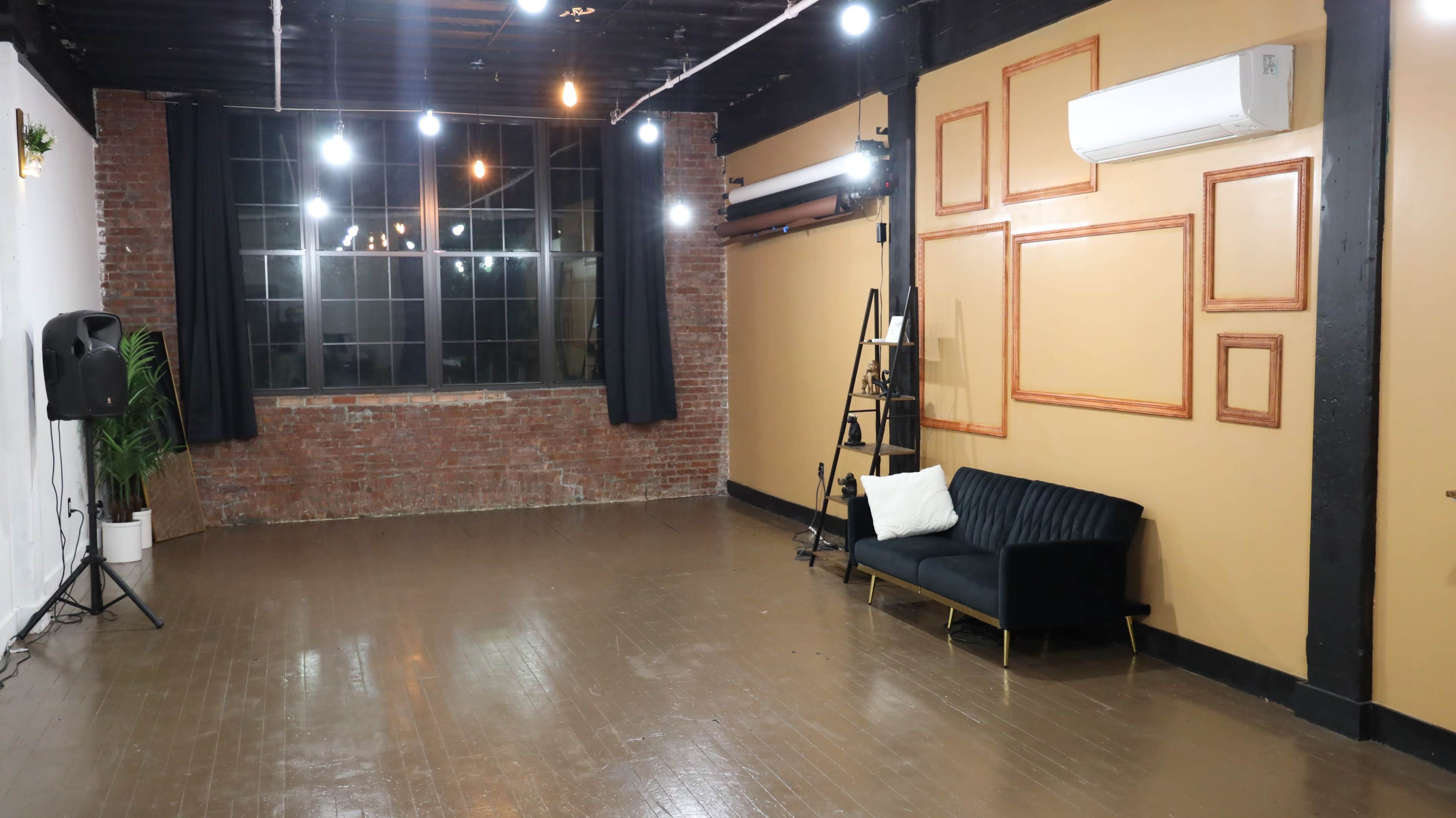An empty room with exposed brick walls, a large window, wooden flooring, and a black sofa against one wall, illuminated by string lights and ceiling fixtures.