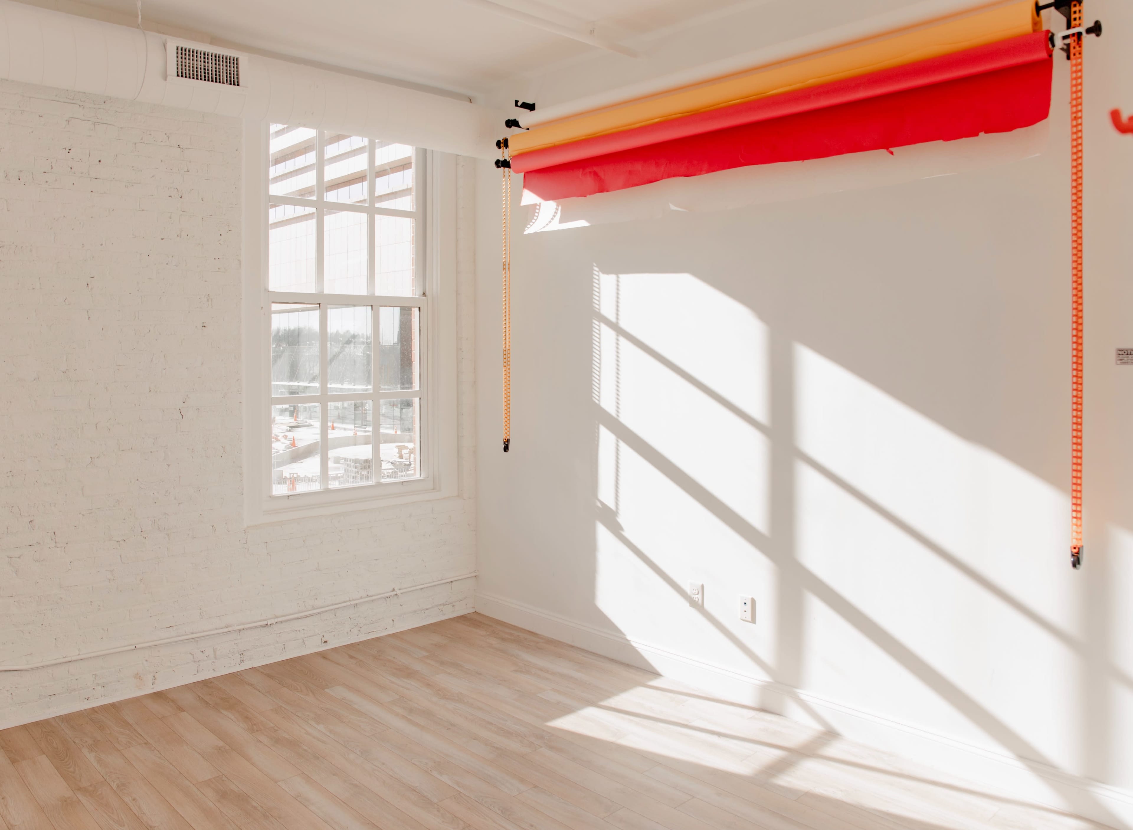 A brightly lit, empty room with white walls and wooden flooring, featuring a large window and colorful backdrop rolls hanging from the ceiling.