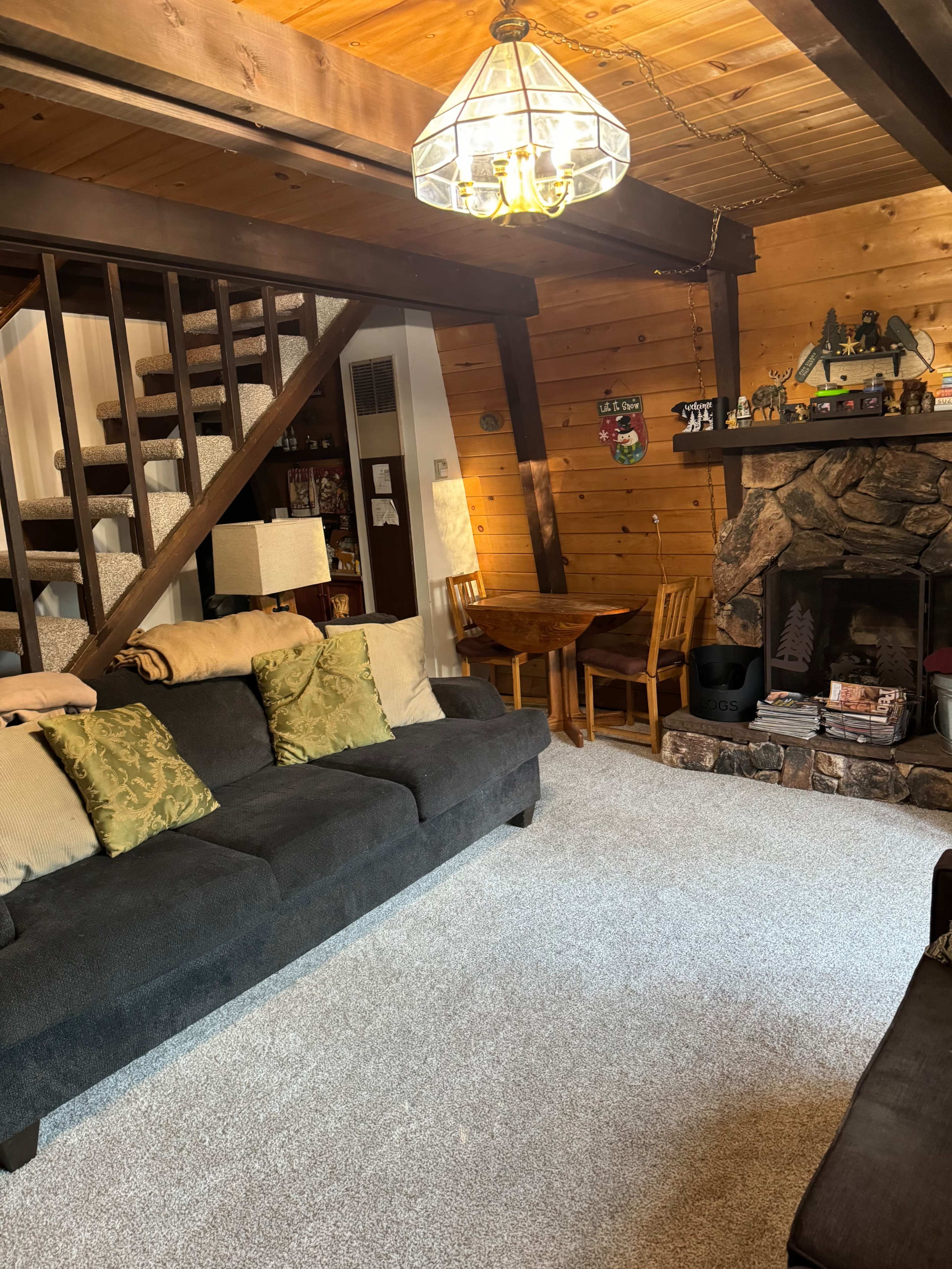 The image shows a cozy living room with a dark sofa, wooden beams, a stone fireplace, and a dining table adjacent to a staircase.