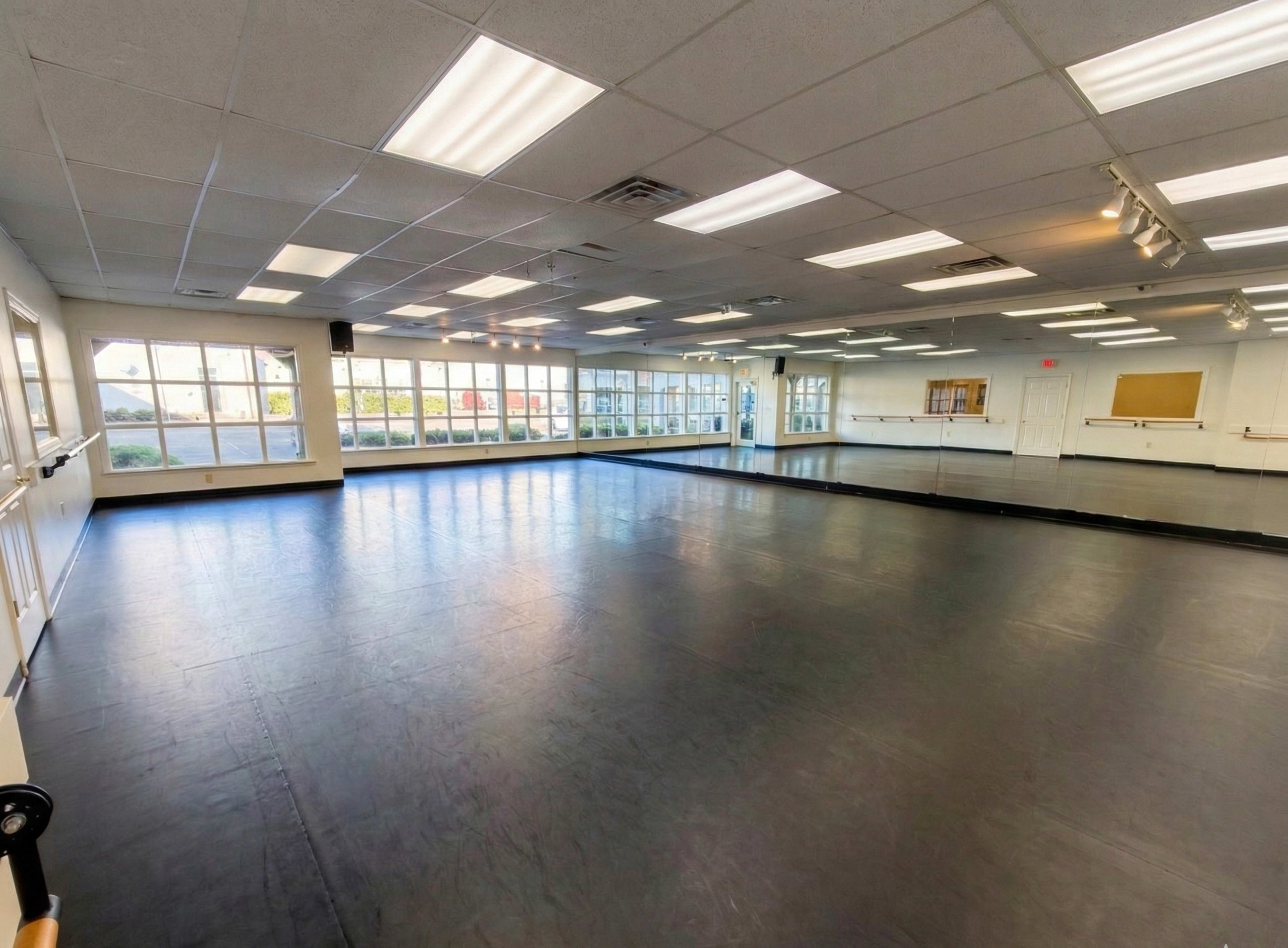 The image shows a spacious dance studio with a mirrored wall, black flooring, and large windows allowing natural light to enter.