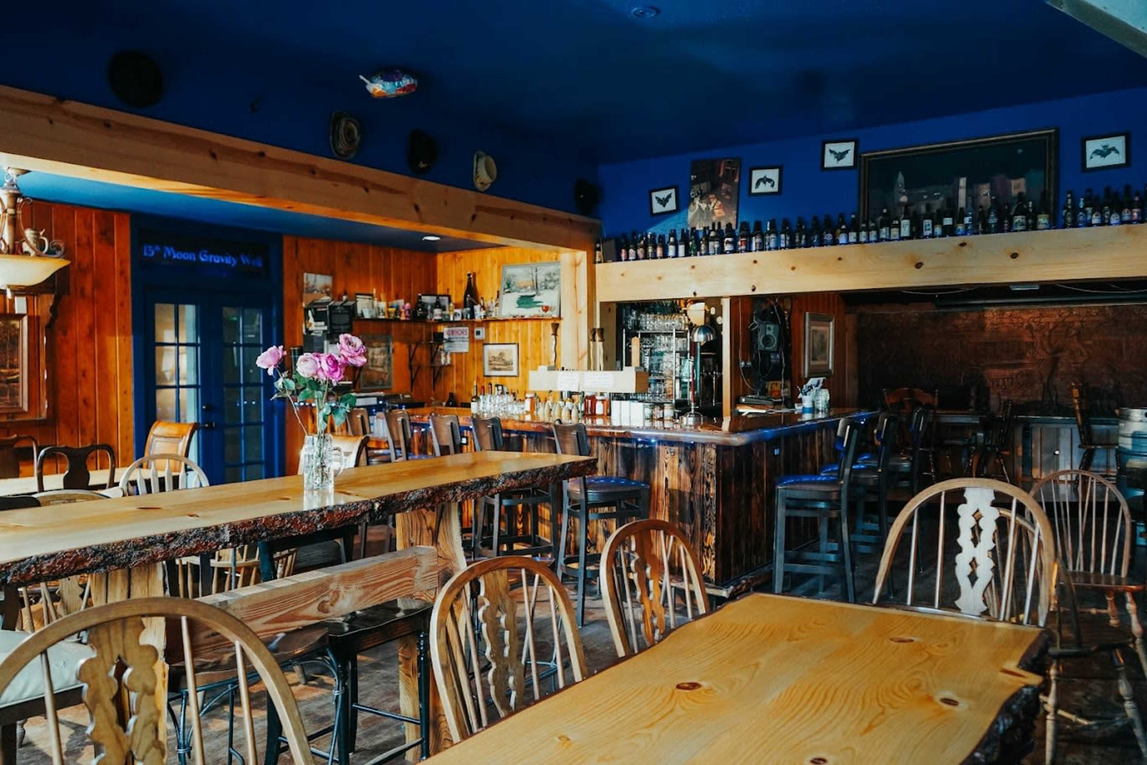 A rustic restaurant interior with wooden tables and chairs, a bar stocked with bottles, and decor including framed pictures and fresh flowers.