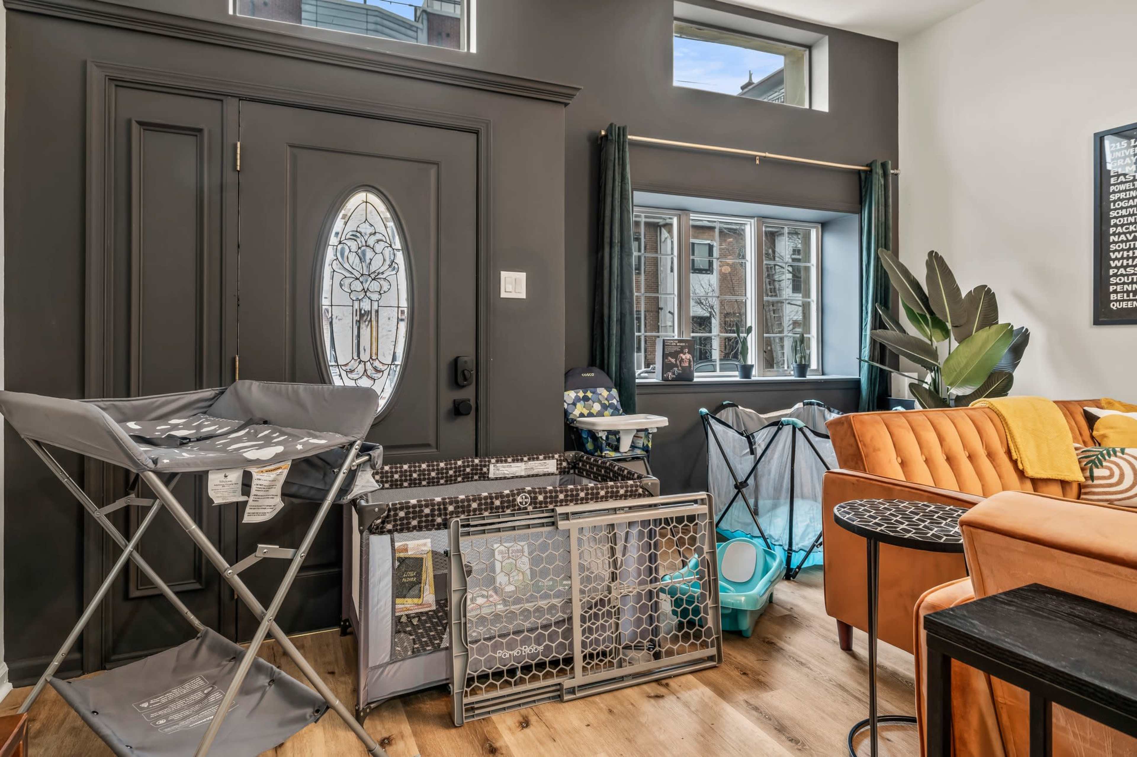 The scene features a dark entryway with a decorative door, a large window, and various baby items like a portable playpen and a foldable chair arranged on the floor.