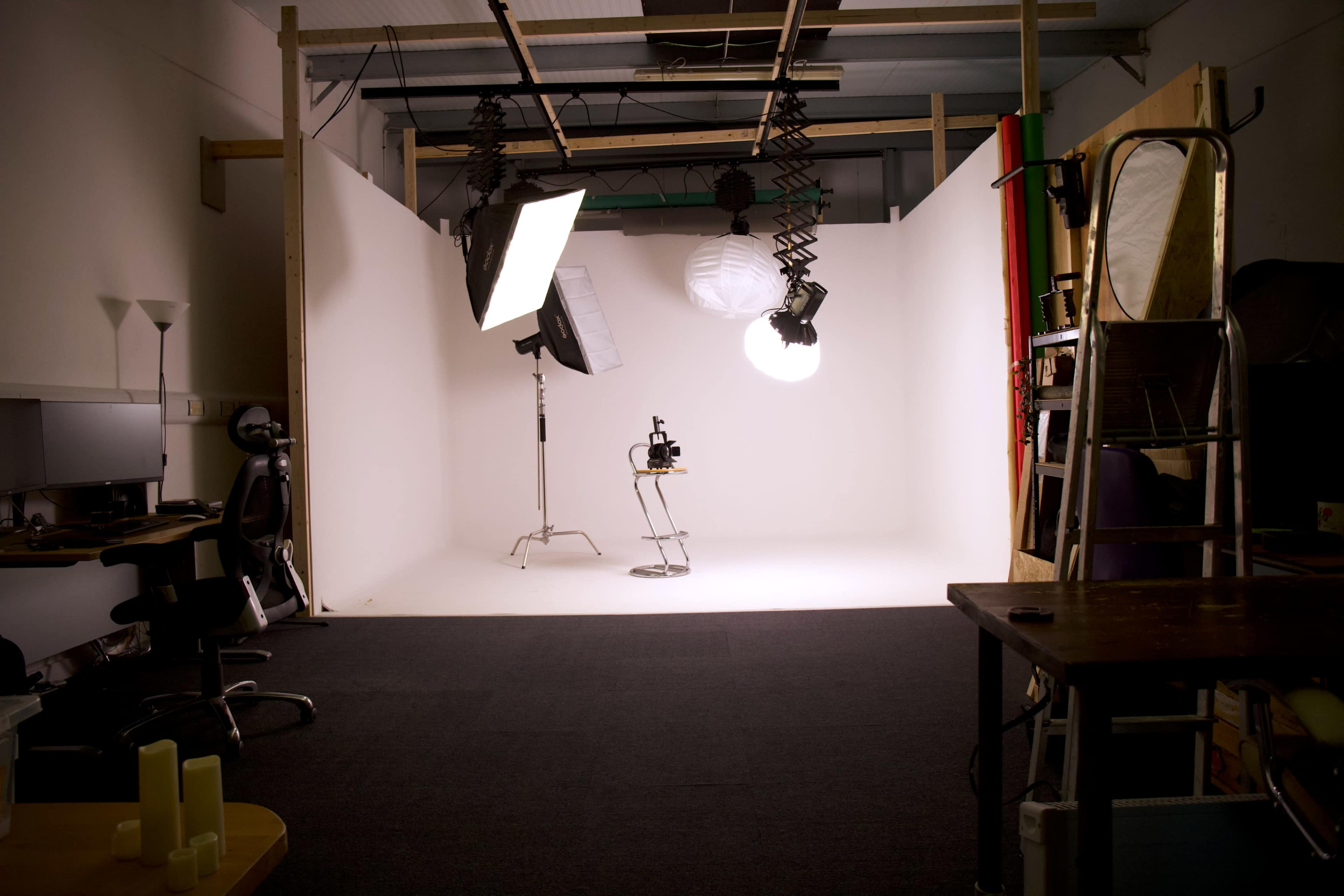 The image depicts a photography studio with a white backdrop, lighting equipment, a chair, and a desk area on one side.