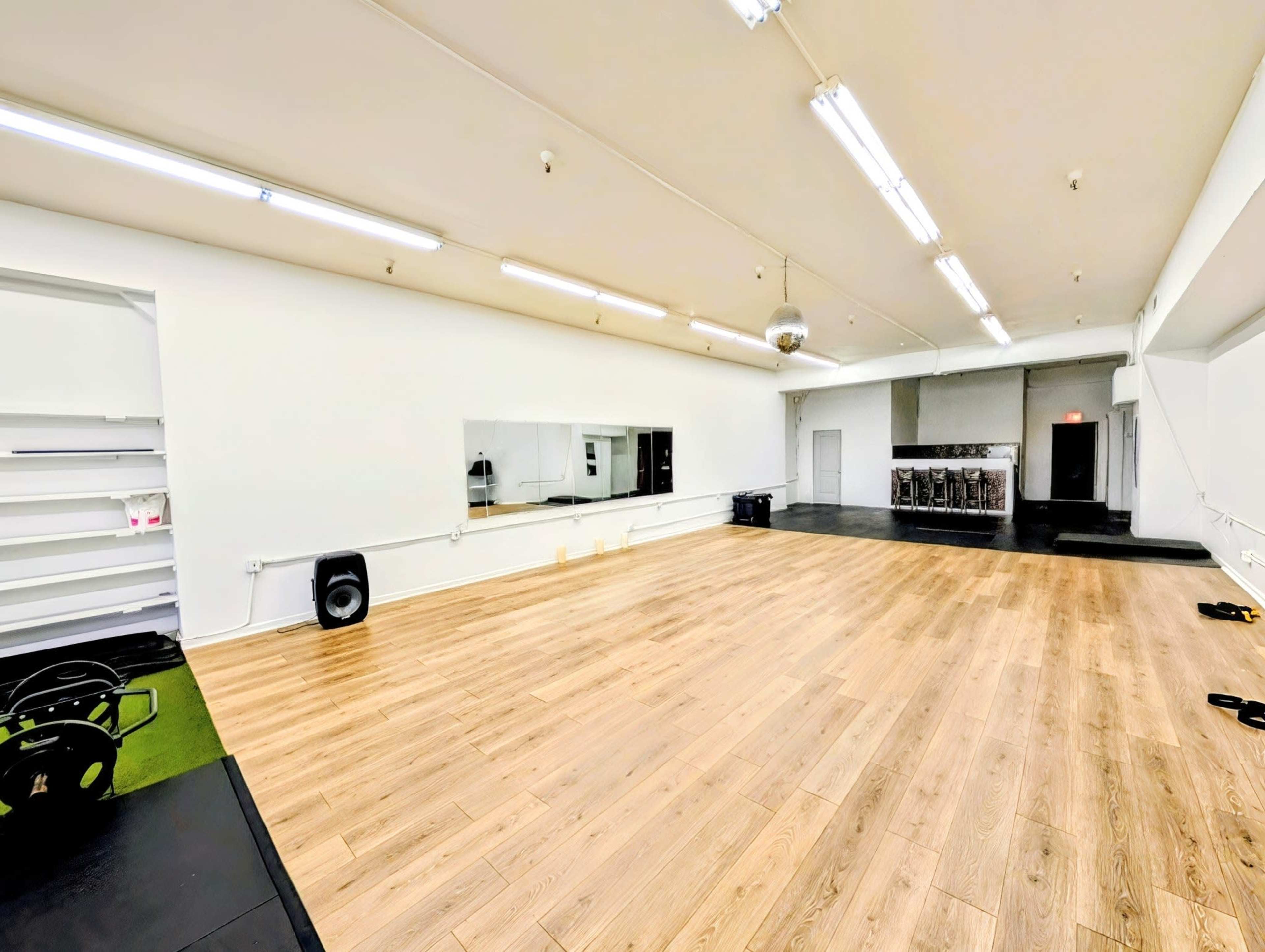 The image shows a spacious, empty fitness studio with wooden flooring, bright overhead lights, a mirrored wall, and a small stage area in the back.