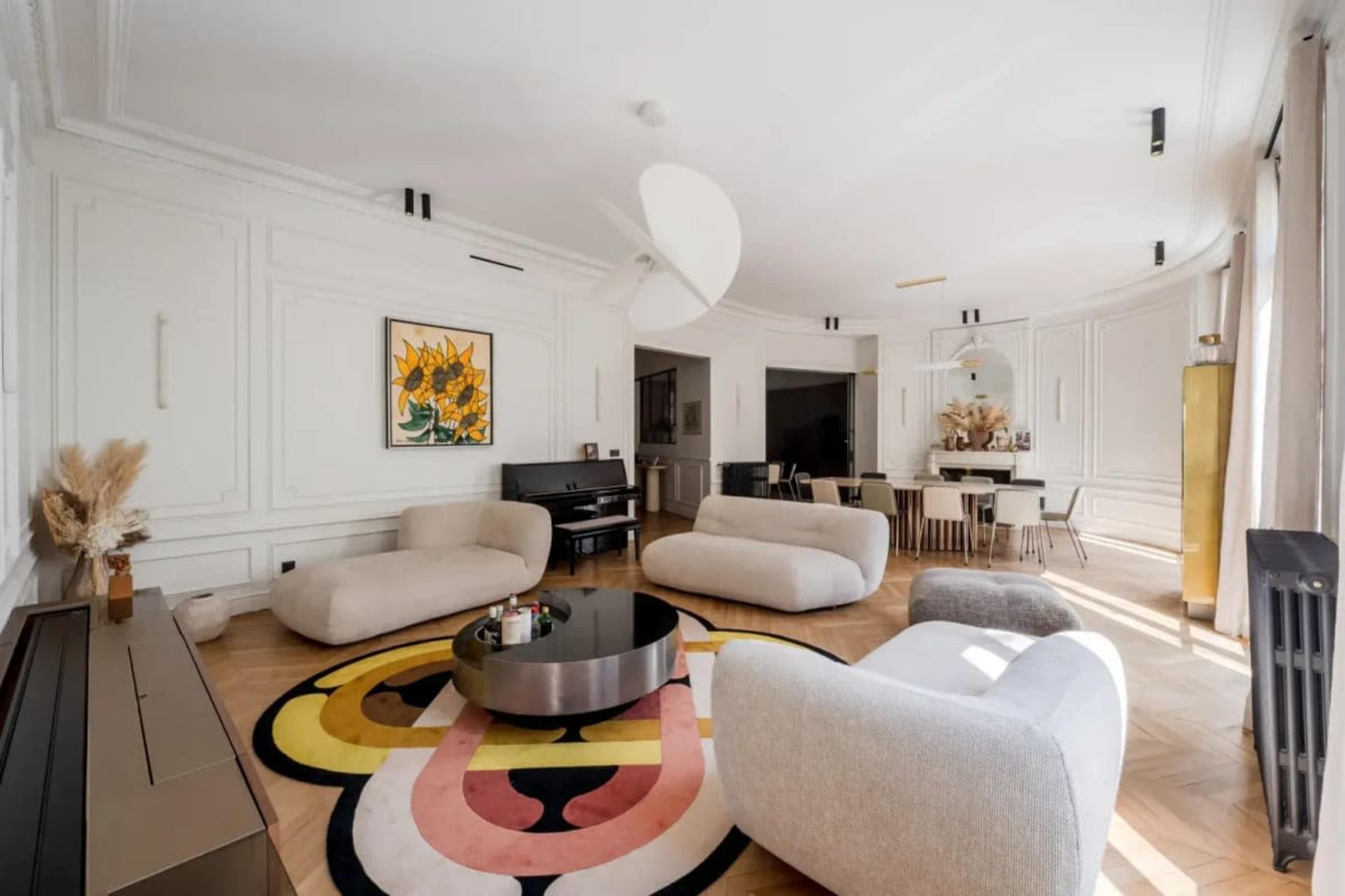The image shows a spacious living room featuring two soft sofas, a round coffee table, and a large, colorful area rug, with stylish decor elements and large windows allowing natural light to flood the space.