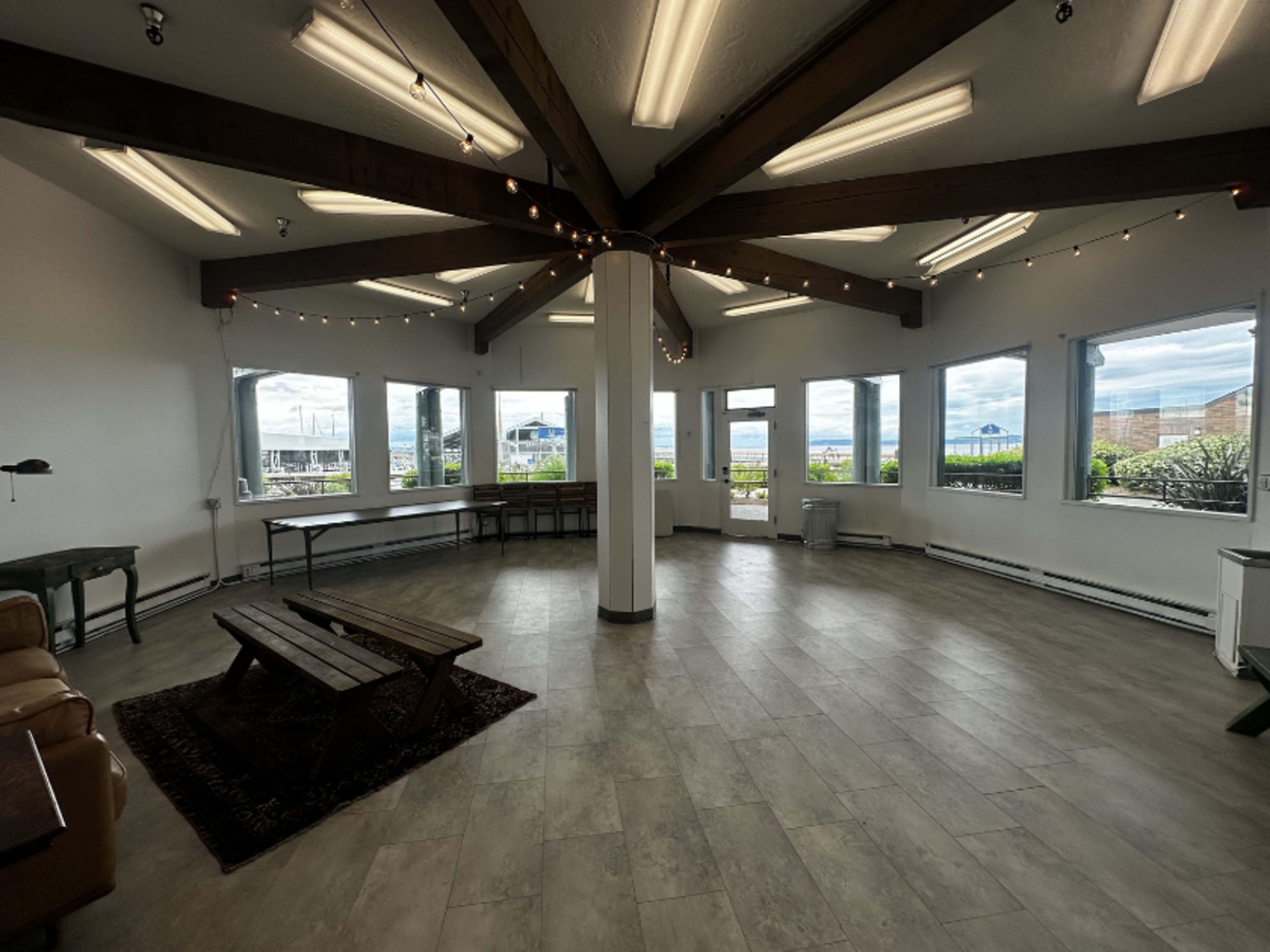 The image shows an empty, spacious room with a circular layout, featuring large windows and a wooden beam ceiling adorned with string lights.
