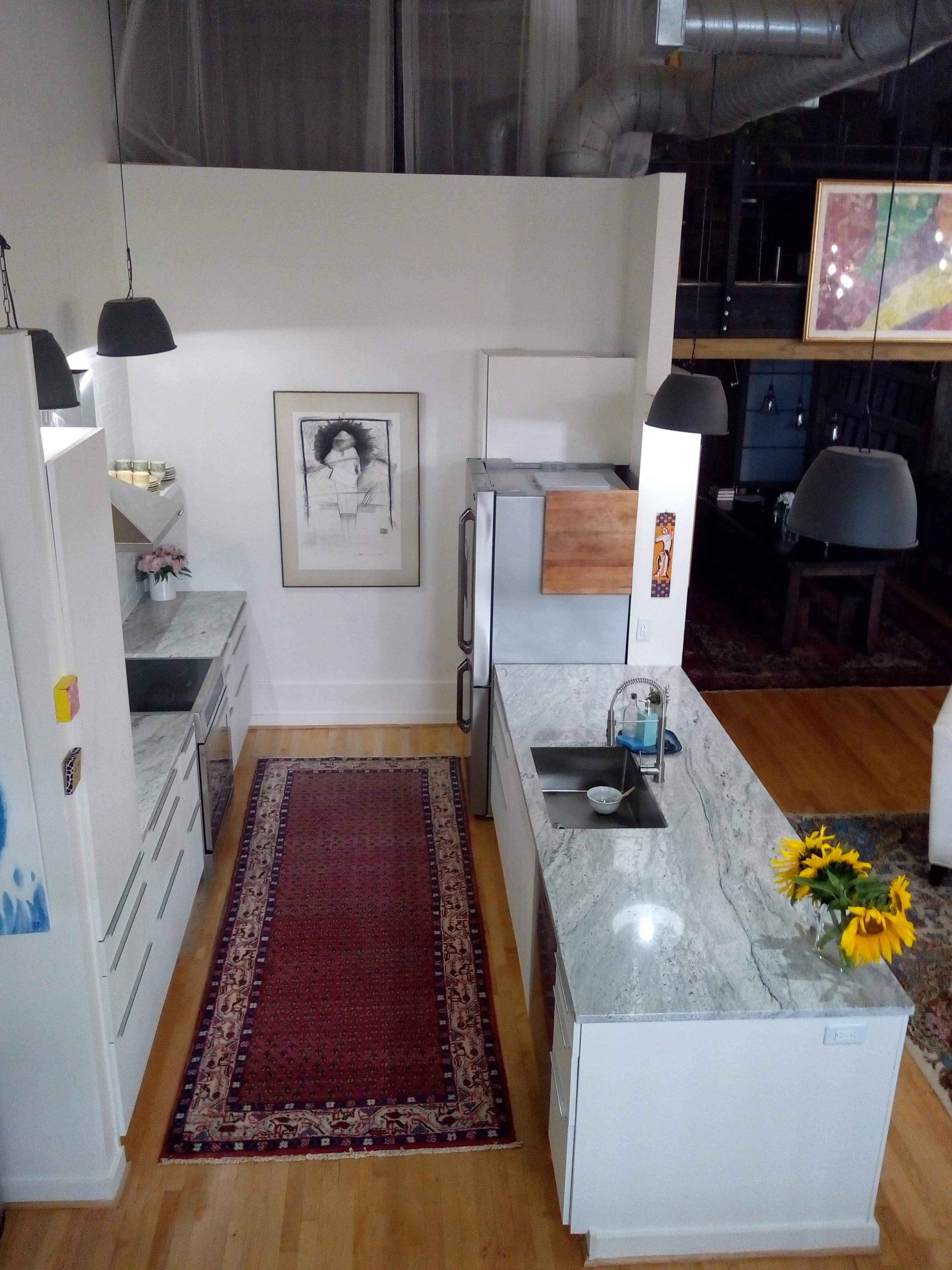 The image shows a modern kitchen with granite countertops, a stainless steel refrigerator, and a colorful rug, featuring a vase of sunflowers on the counter.