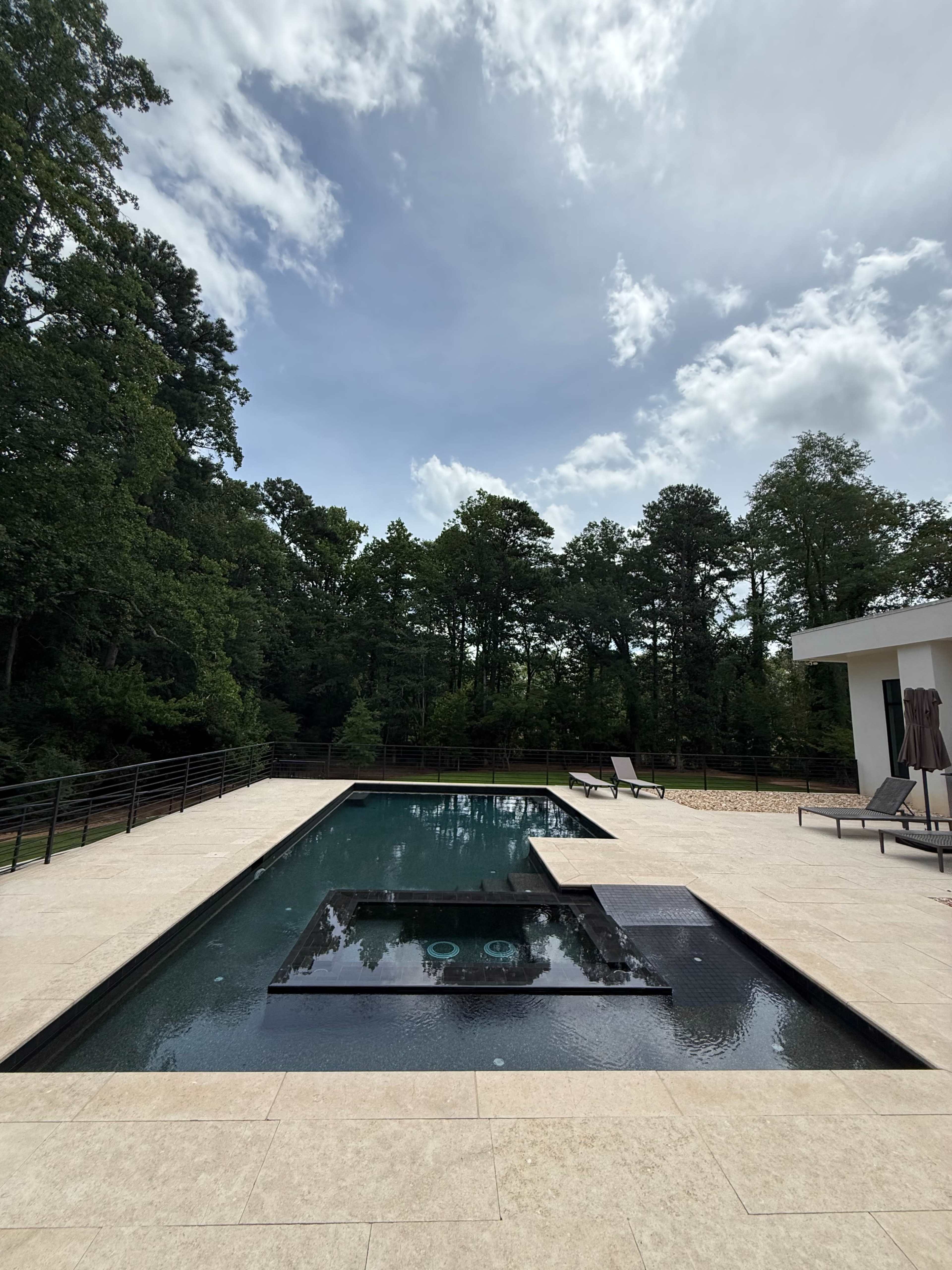 The image shows a modern swimming pool surrounded by a stone deck, with lounge chairs and trees in the background under a partly cloudy sky.