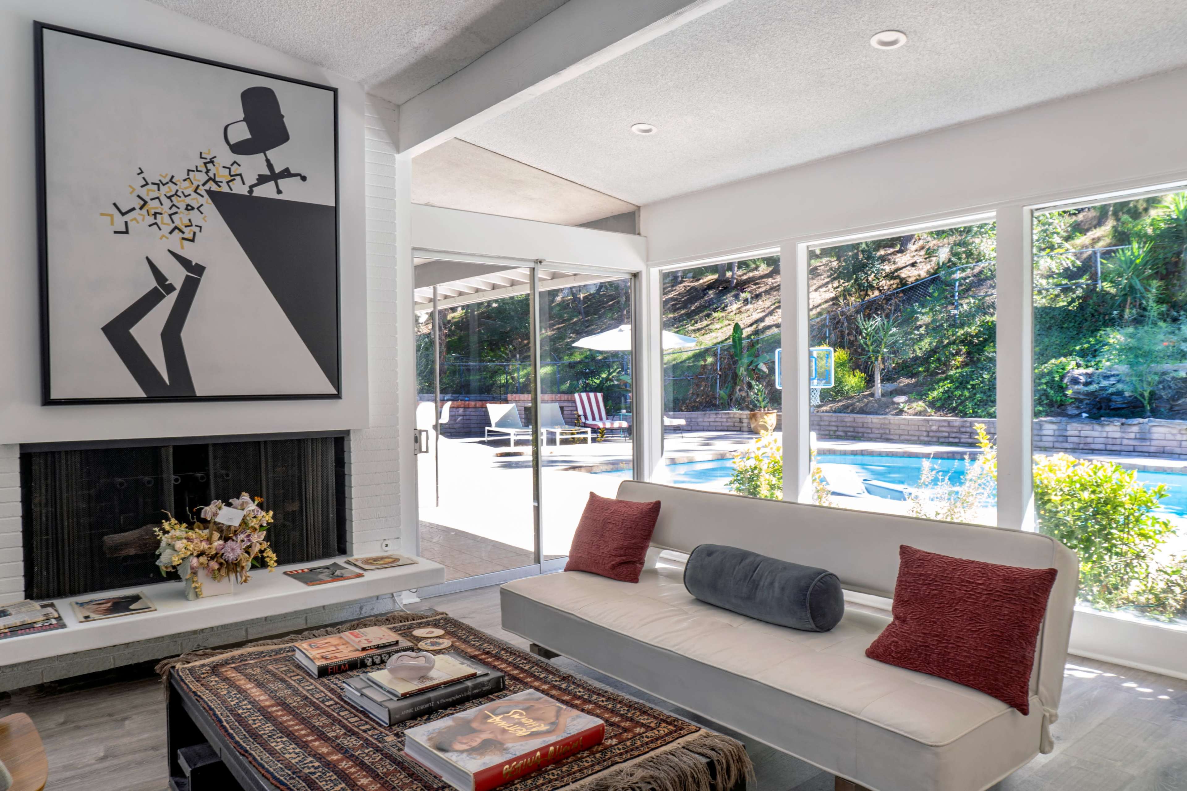 A modern living room features a white sofa, a large abstract artwork on the wall, and large windows overlooking a swimming pool surrounded by greenery.
