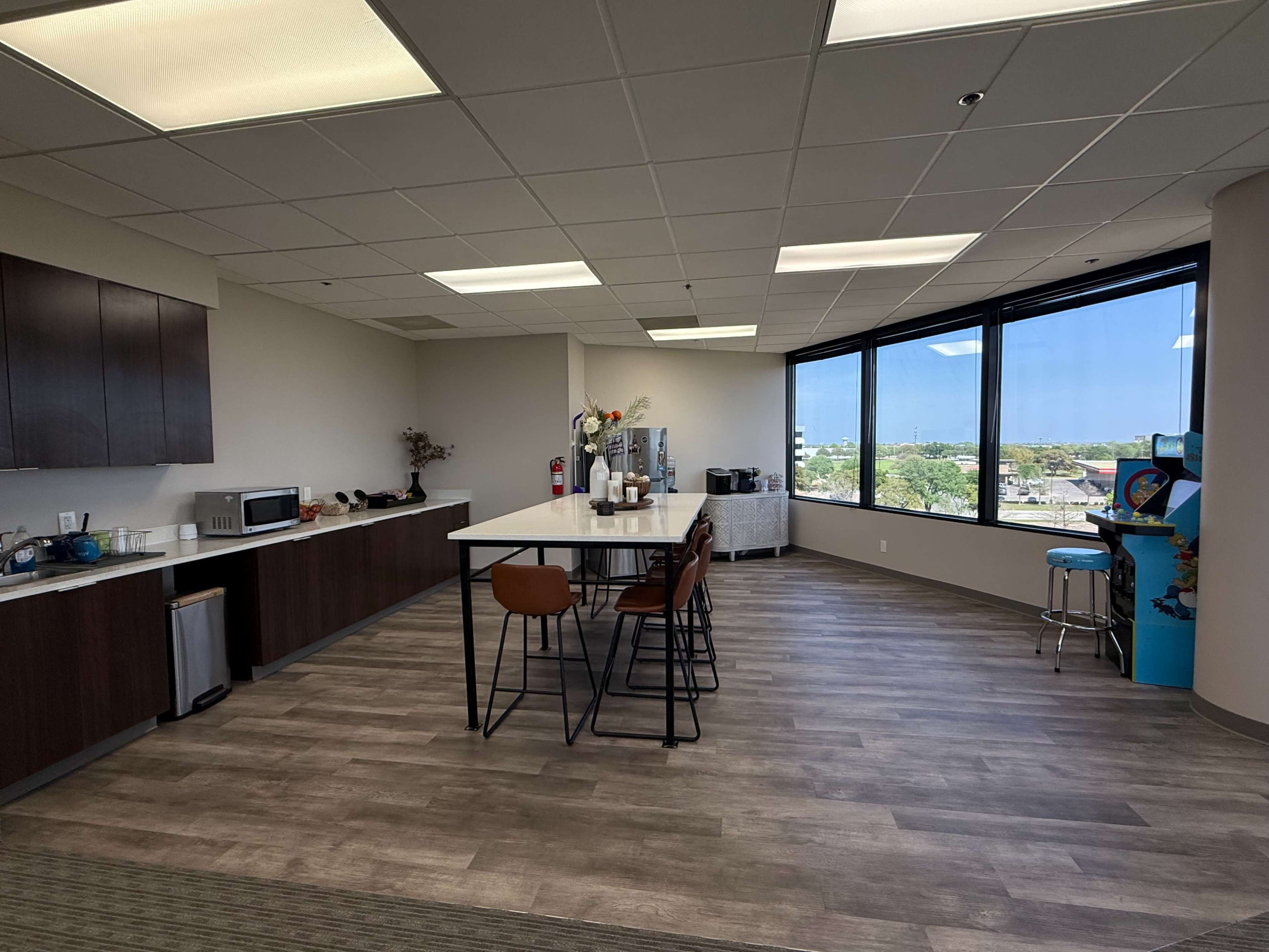 The image depicts a modern break room with a kitchenette, a long table with seating, and large windows offering a view of the outdoors.