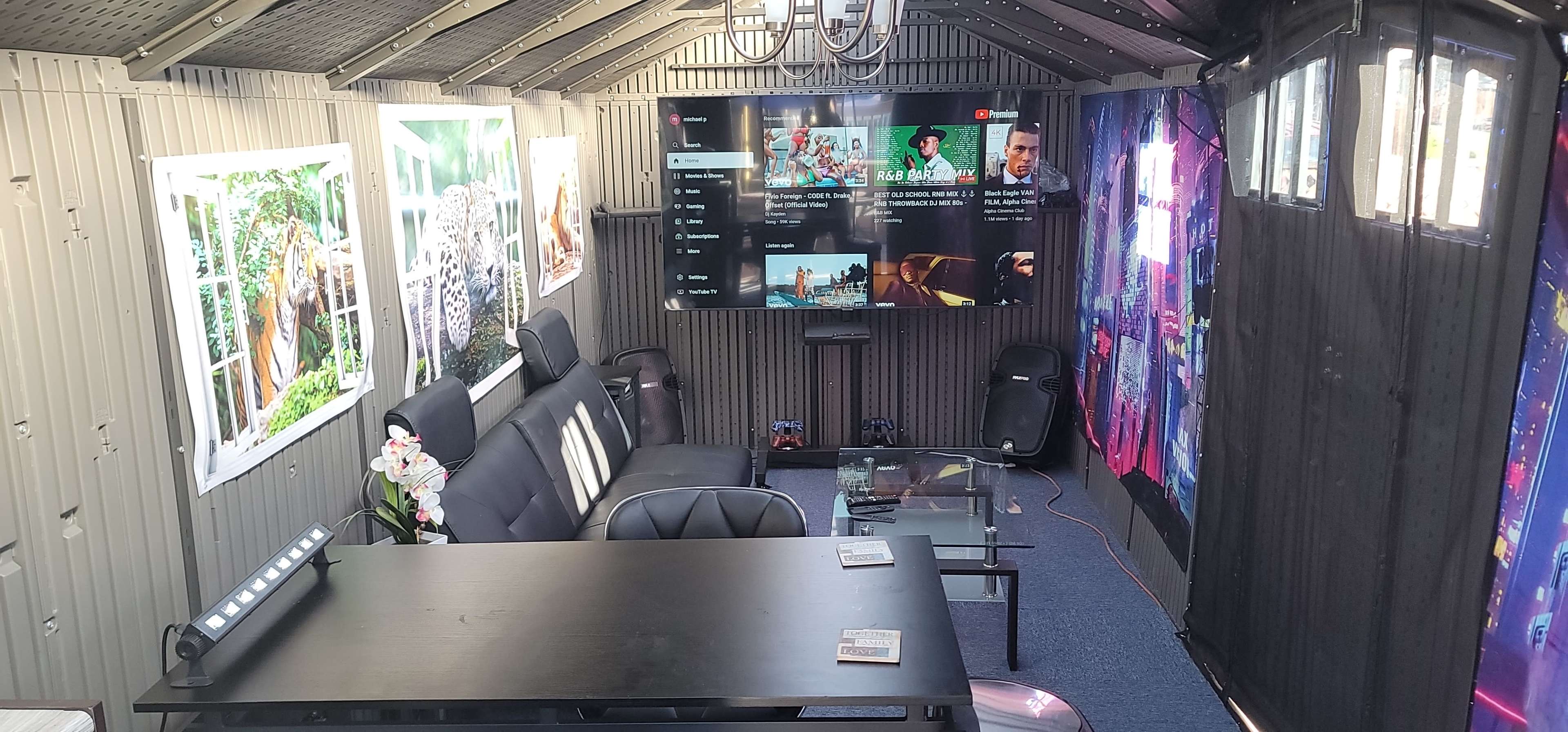 The interior of a shed-style room features a seating area with a black sofa, a large TV mounted on the wall, and decorative wall panels.