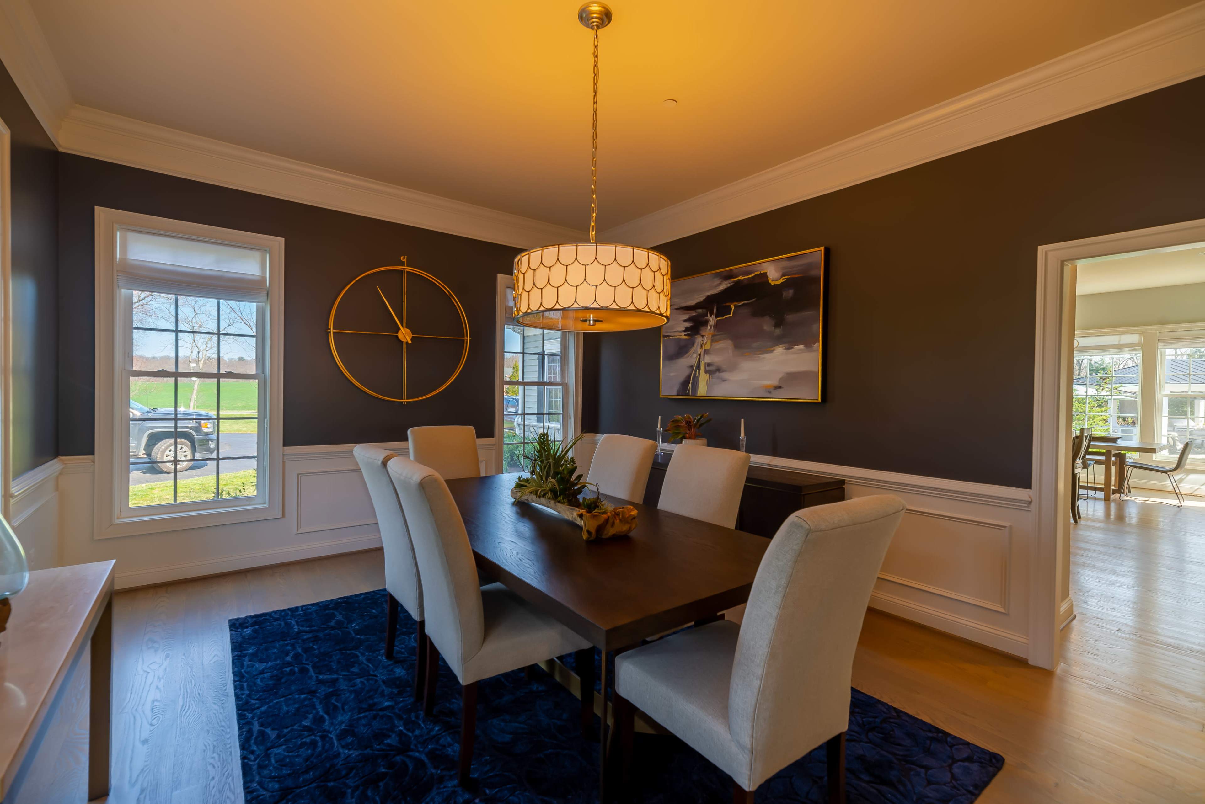 A dining room features a rectangular table surrounded by six upholstered chairs, a large chandelier, and framed artwork against dark walls, with windows allowing natural light.