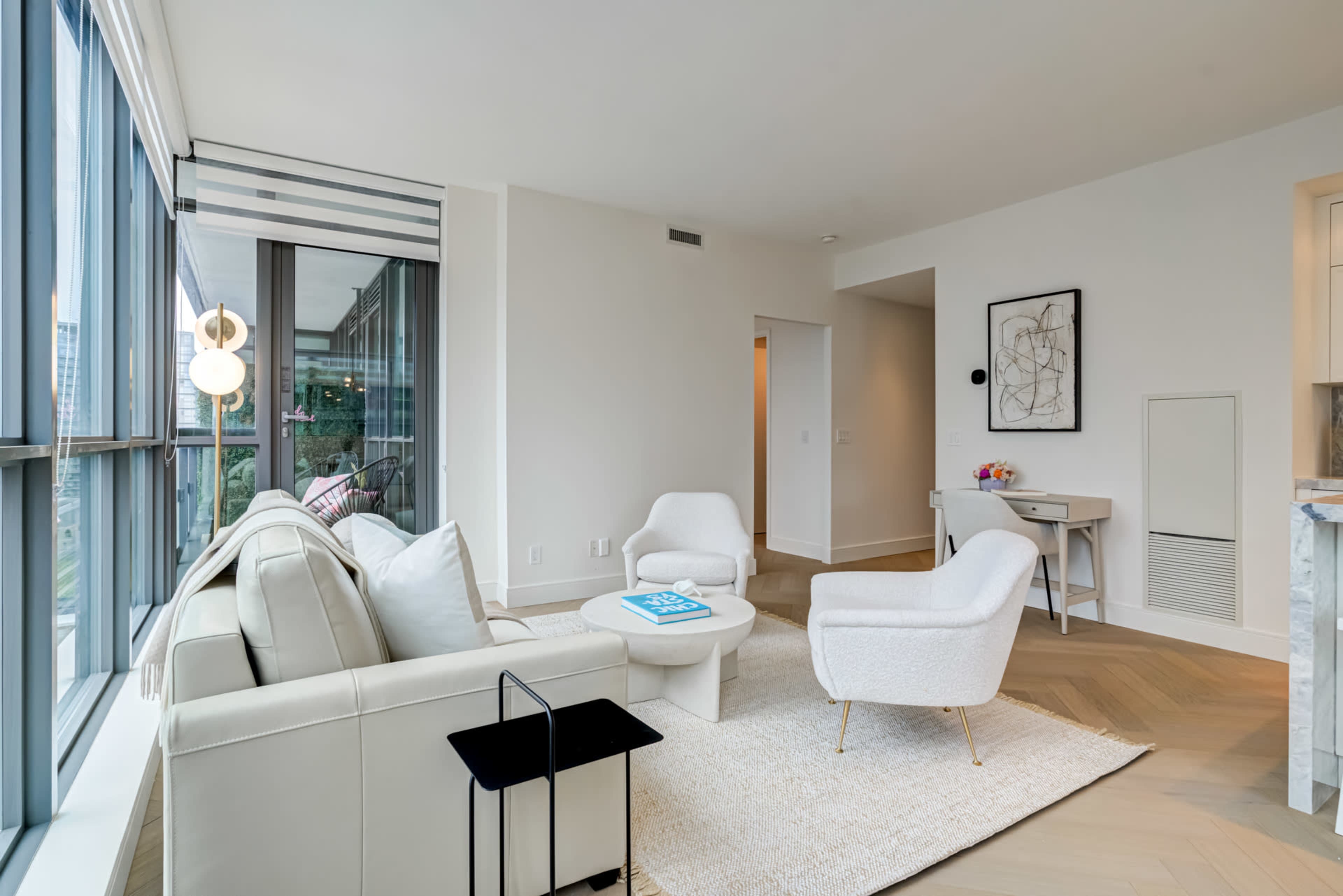 A modern living room with large windows, featuring a light-colored sofa, two white chairs, a round coffee table, and minimalistic decor.