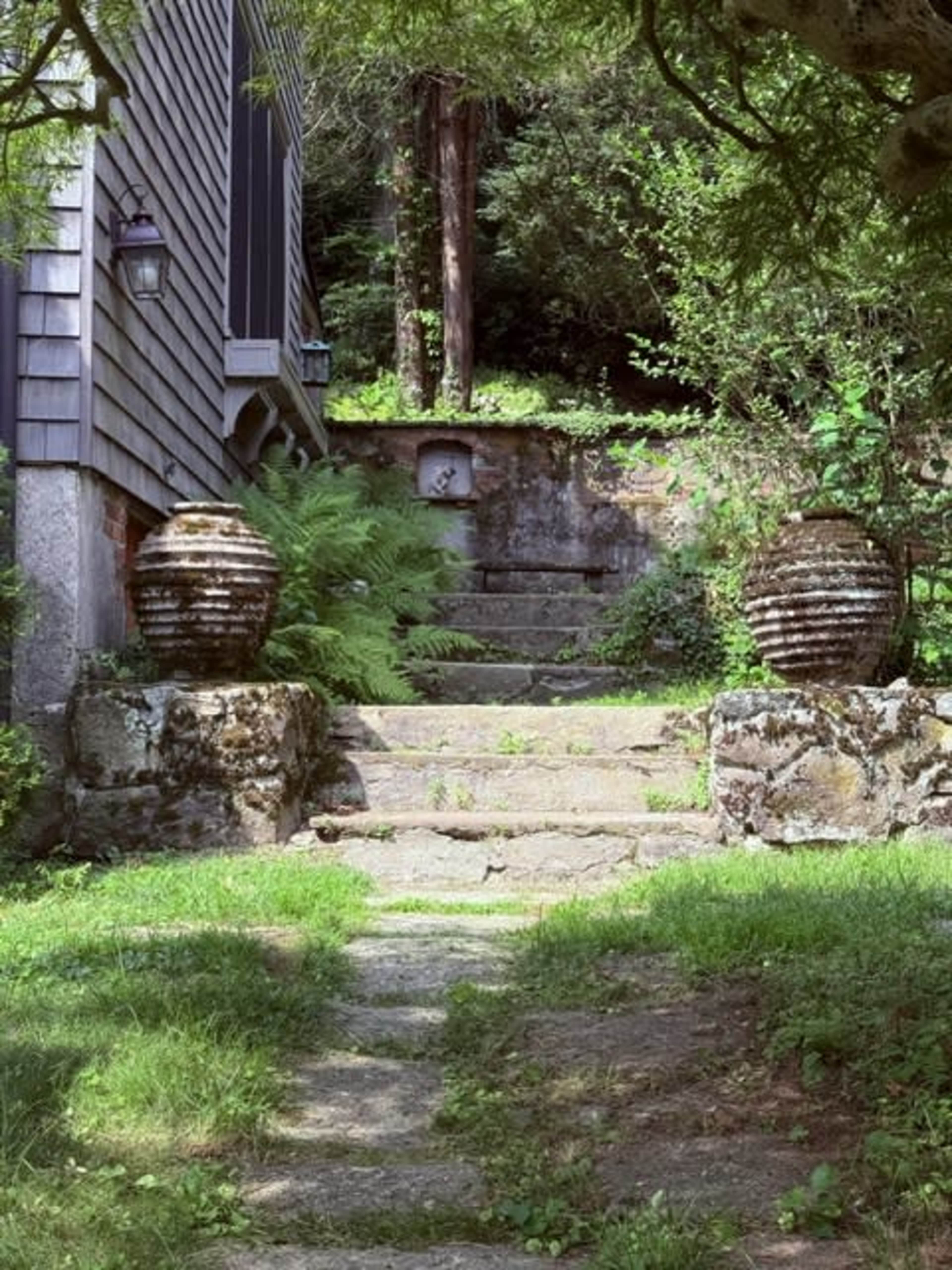 The image shows a stone pathway leading up to a wooded area, flanked by two decorative urns on either side.
