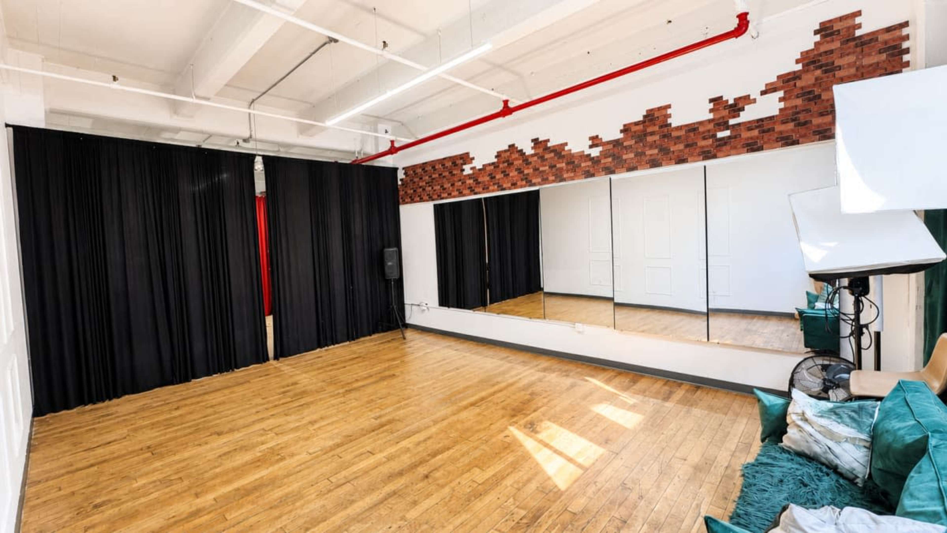 The image shows a spacious studio with wooden flooring, large mirrors on one wall, black drapes, and minimal furnishings.