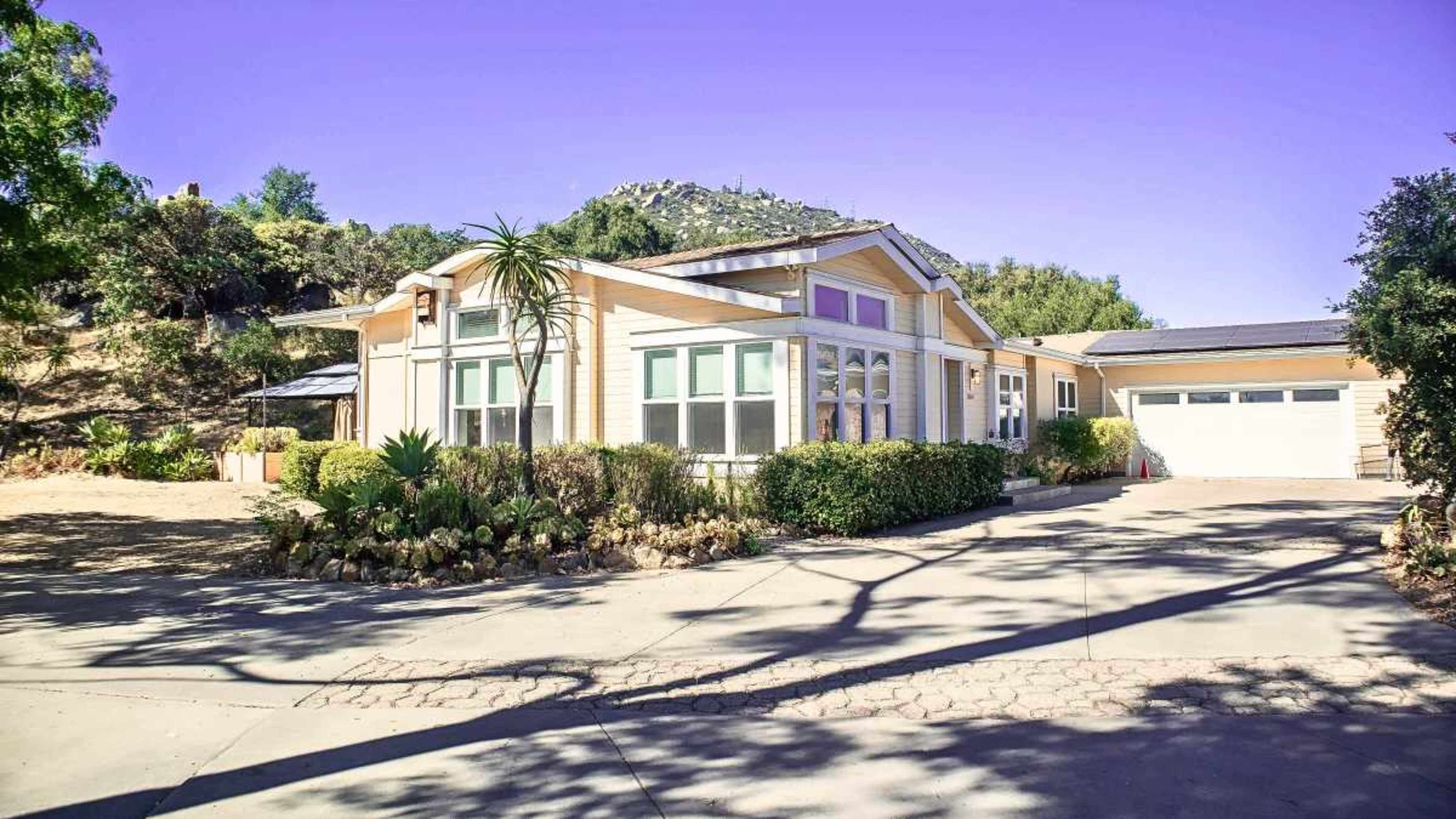 The image shows a large, single-story house with a landscaped front yard, a driveway leading to a garage, and a mountainous background.