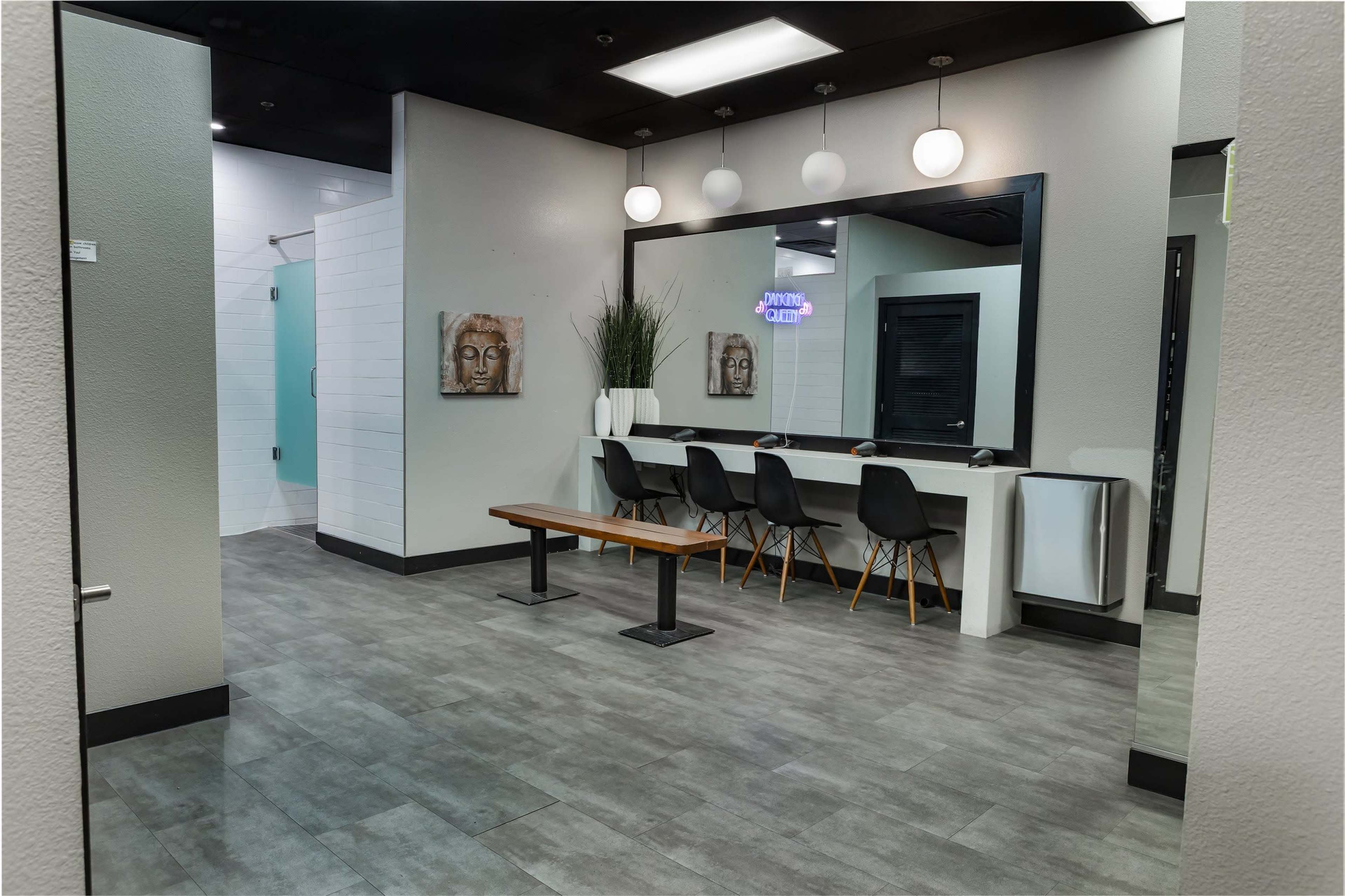 The image depicts a modern, well-lit lobby area with a large mirror, a wooden bench, several black chairs, and a partition leading to a shower space.