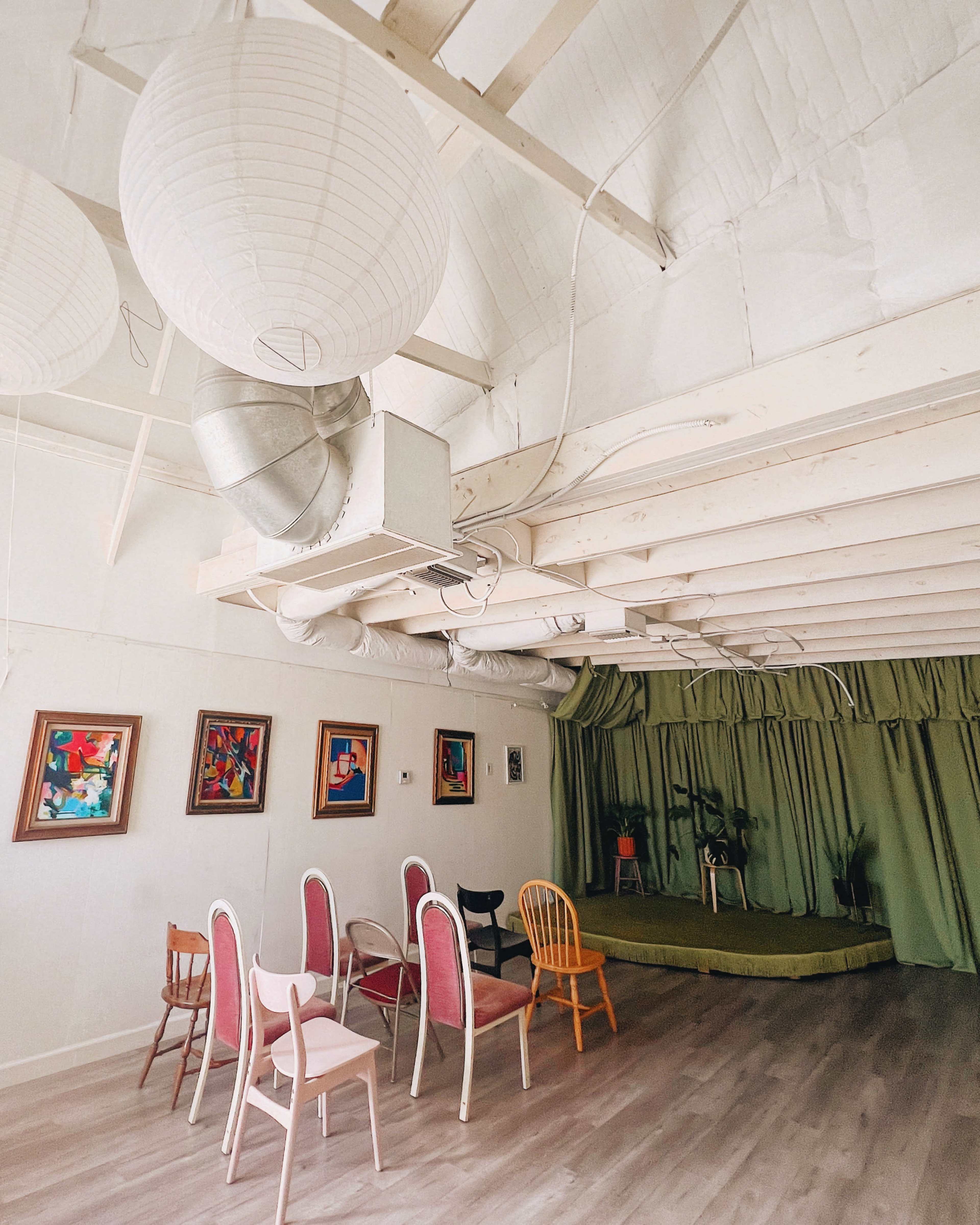 The image shows a spacious room with a wooden floor, adorned with several colorful artworks, a green curtain backdrop, and various chairs arranged around.