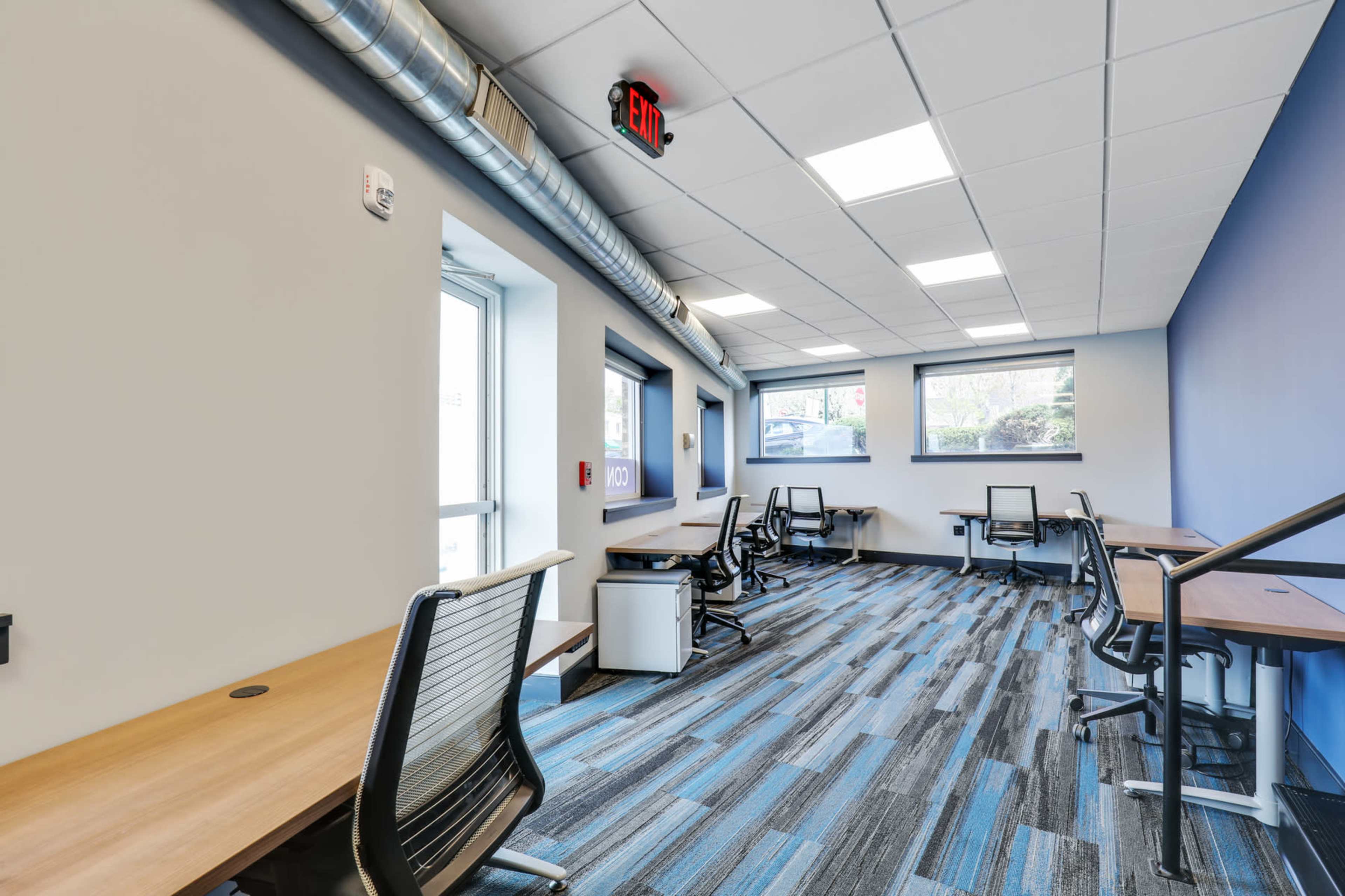 The image depicts a modern, vacant office space with workstations and large windows, featuring wooden desks and ergonomic chairs.
