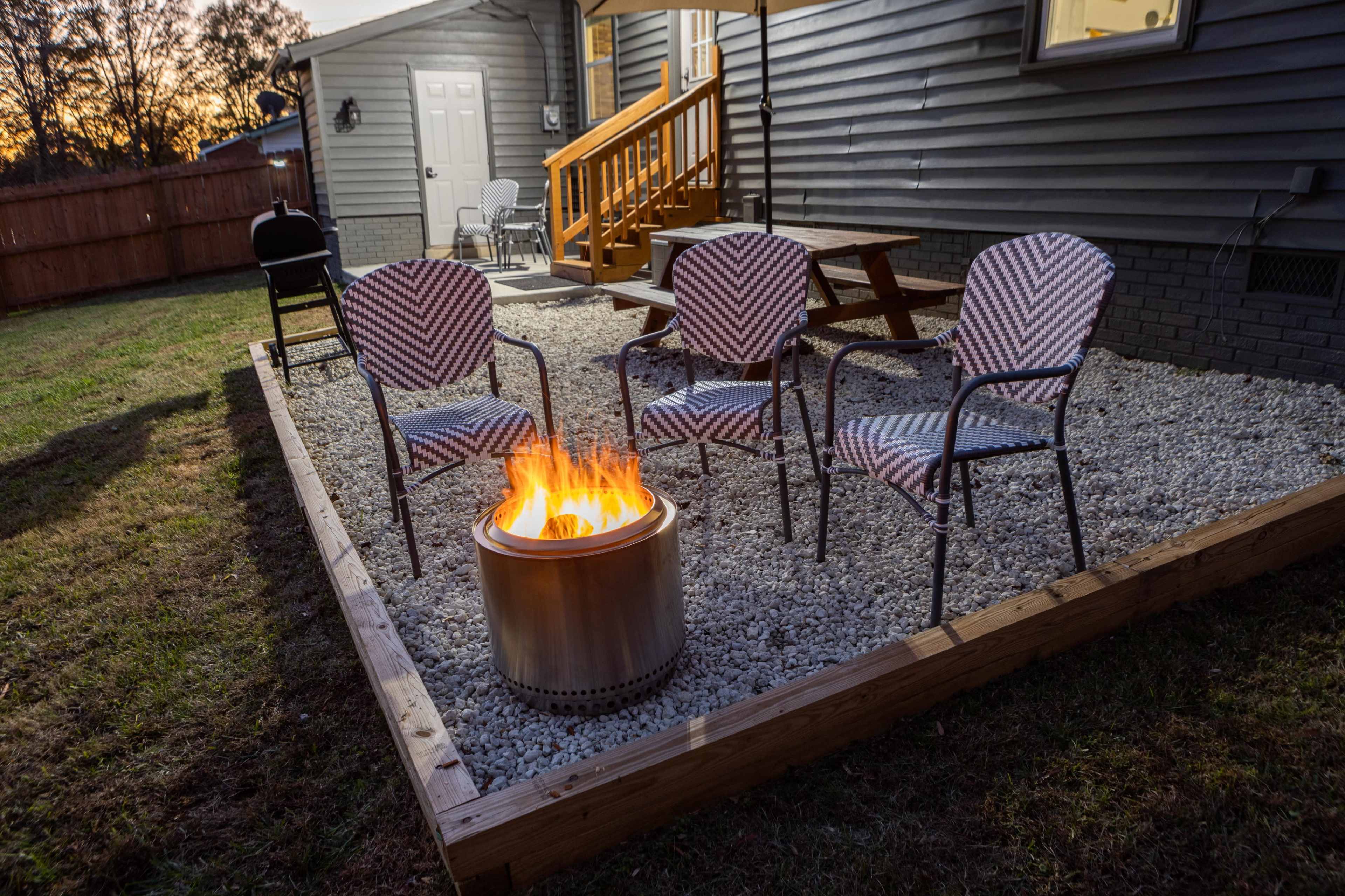 A fire pit surrounded by four chairs is set up on a gravel patio next to a house at dusk.