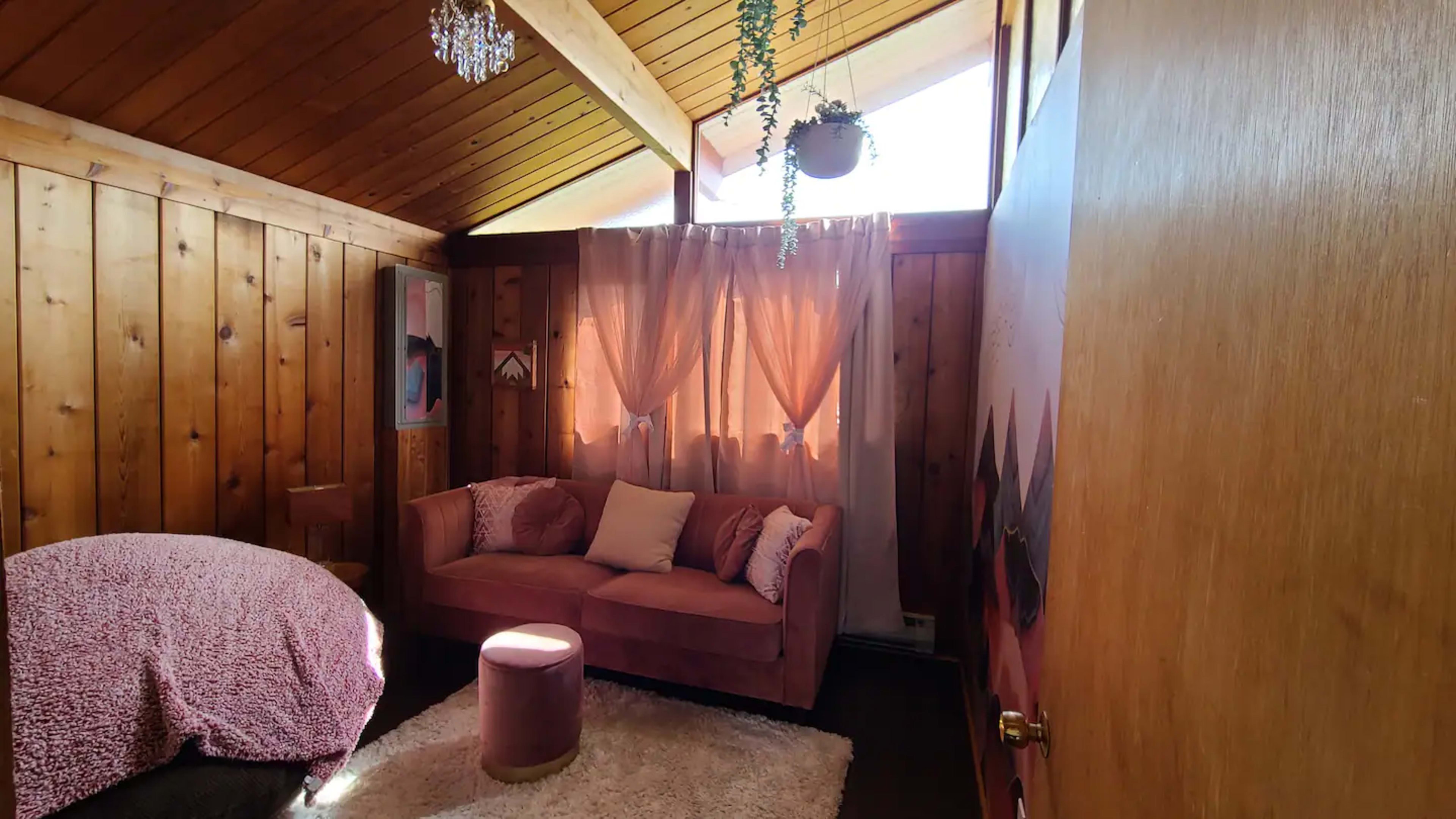 The room features wooden walls, a pink couch, a small round ottoman, and large windows with sheer curtains allowing natural light to enter.