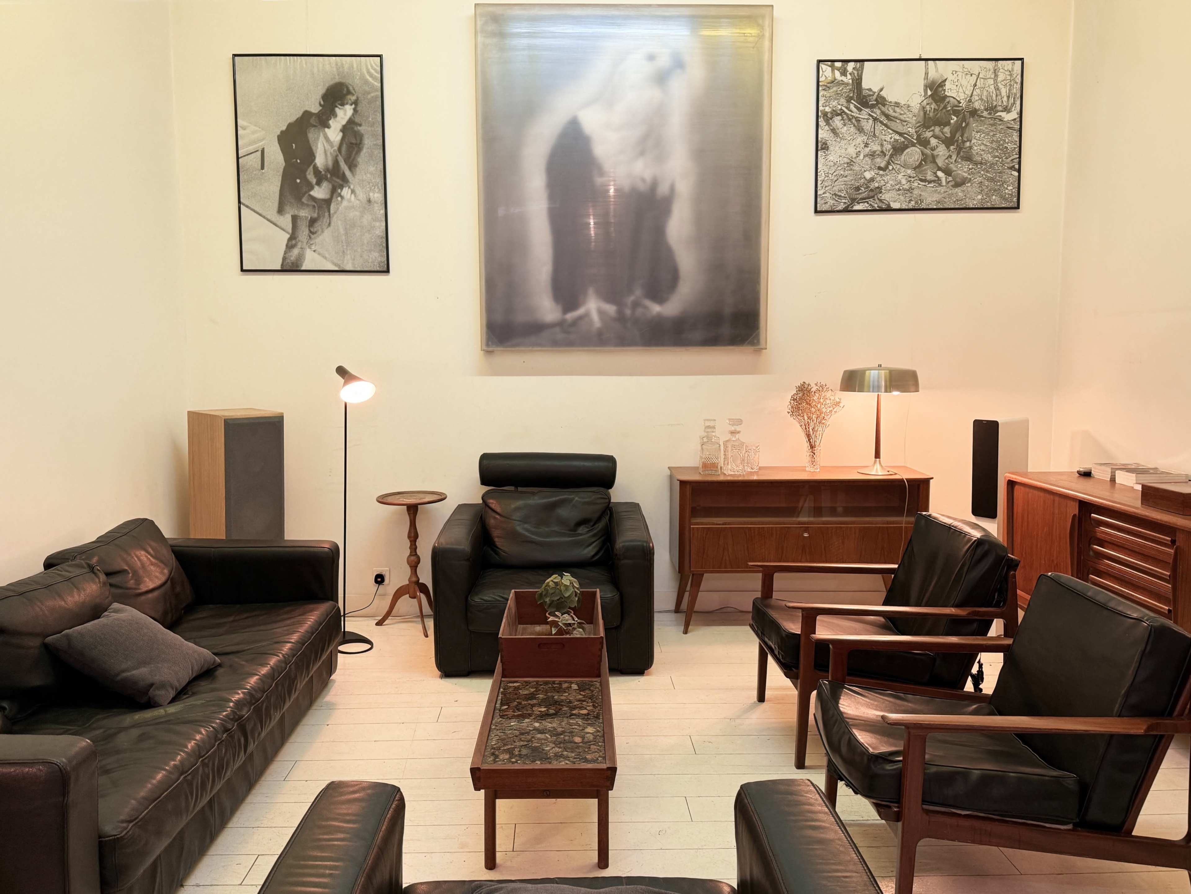The scene features a cozy interior with black leather sofas, wooden chairs, a coffee table, and framed black-and-white photographs on the walls.