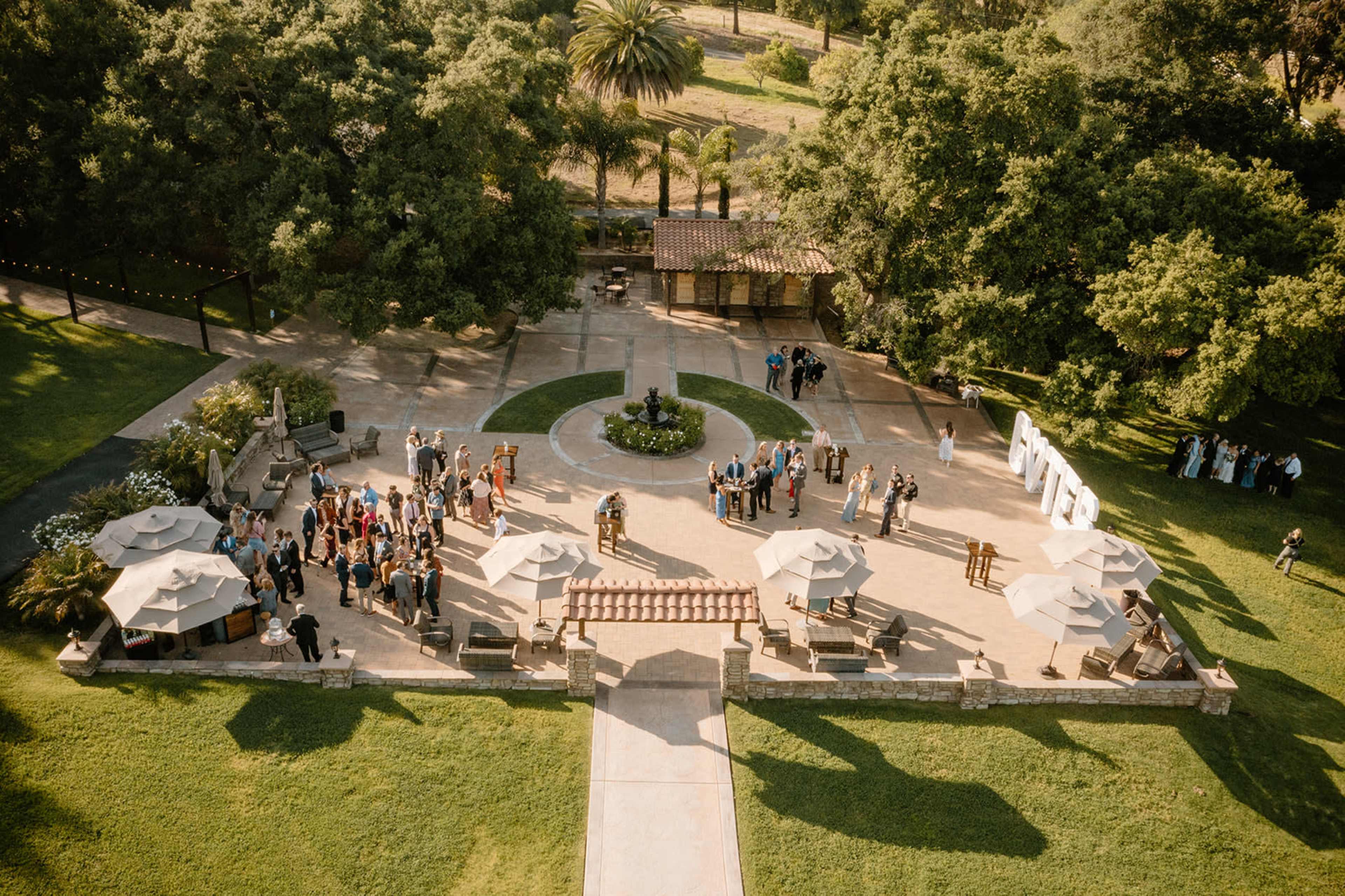 Aerial view of a landscaped outdoor venue with a central circular garden, surrounded by guests mingling under umbrellas and trees.