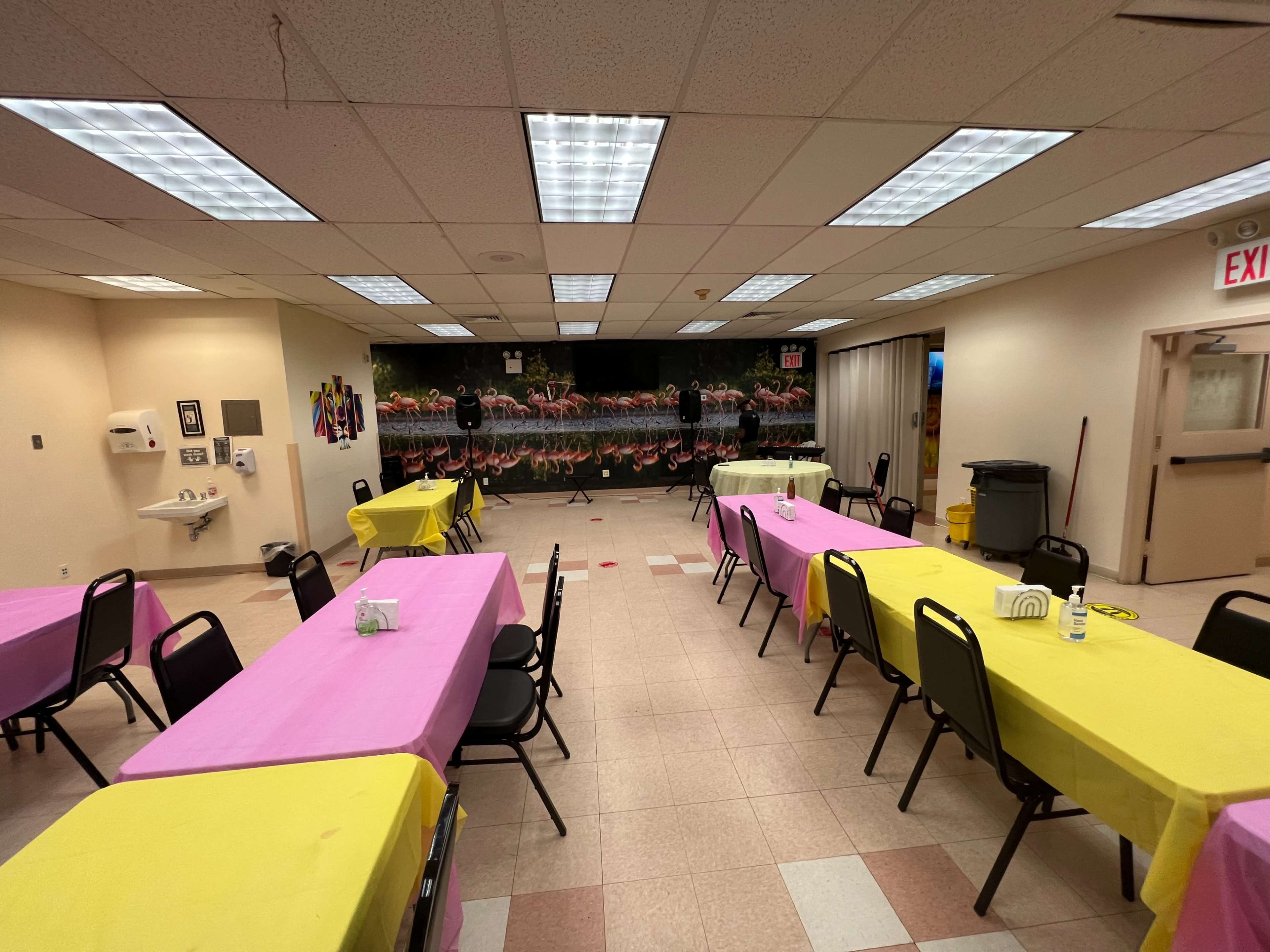 A large room set up for an event, featuring tables covered in pink and yellow tablecloths with a decorative mural on the back wall.