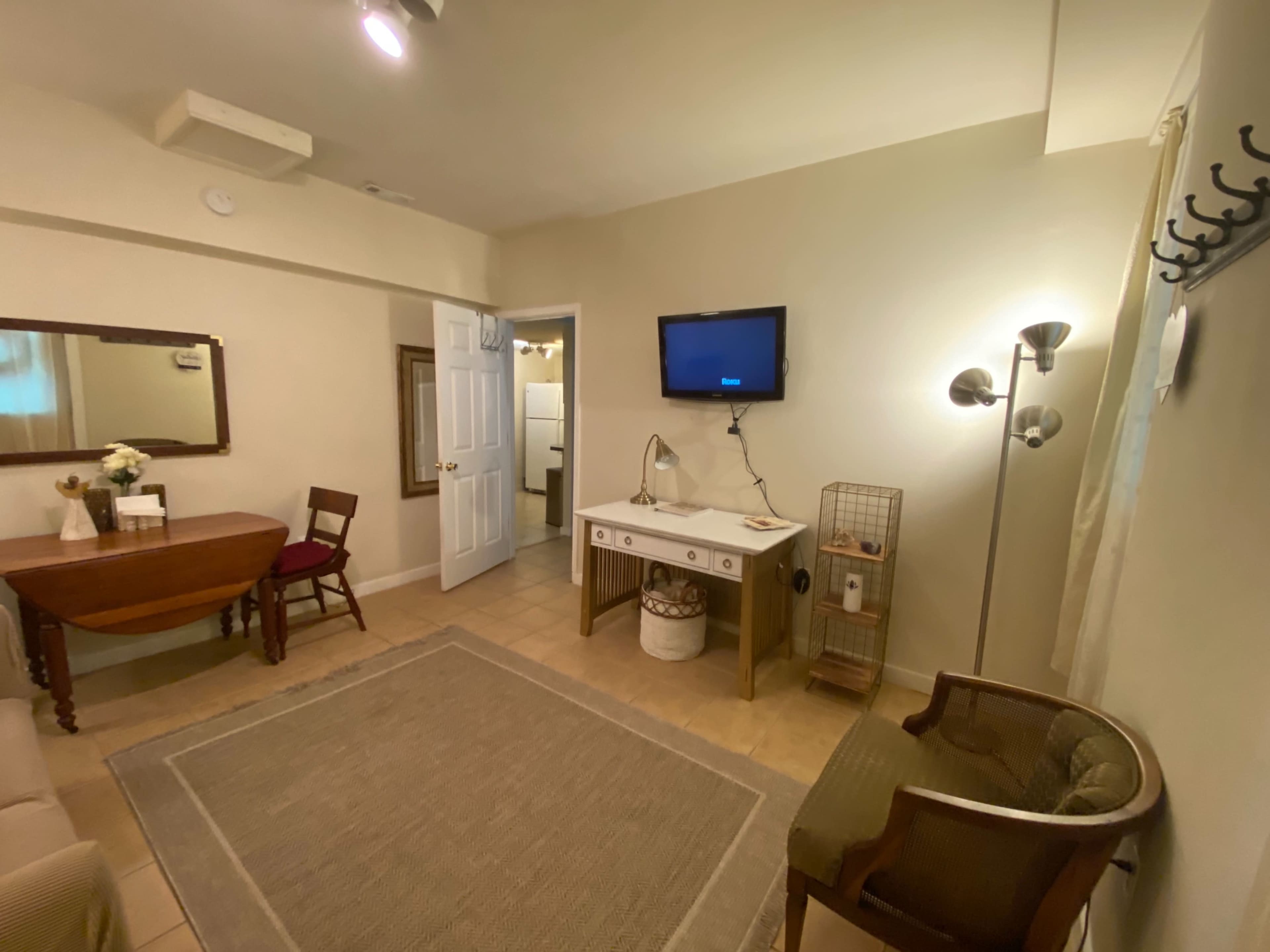 The image shows a small room furnished with a table, a chair, a mirror, a television mounted on the wall, and a rug on the tiled floor.